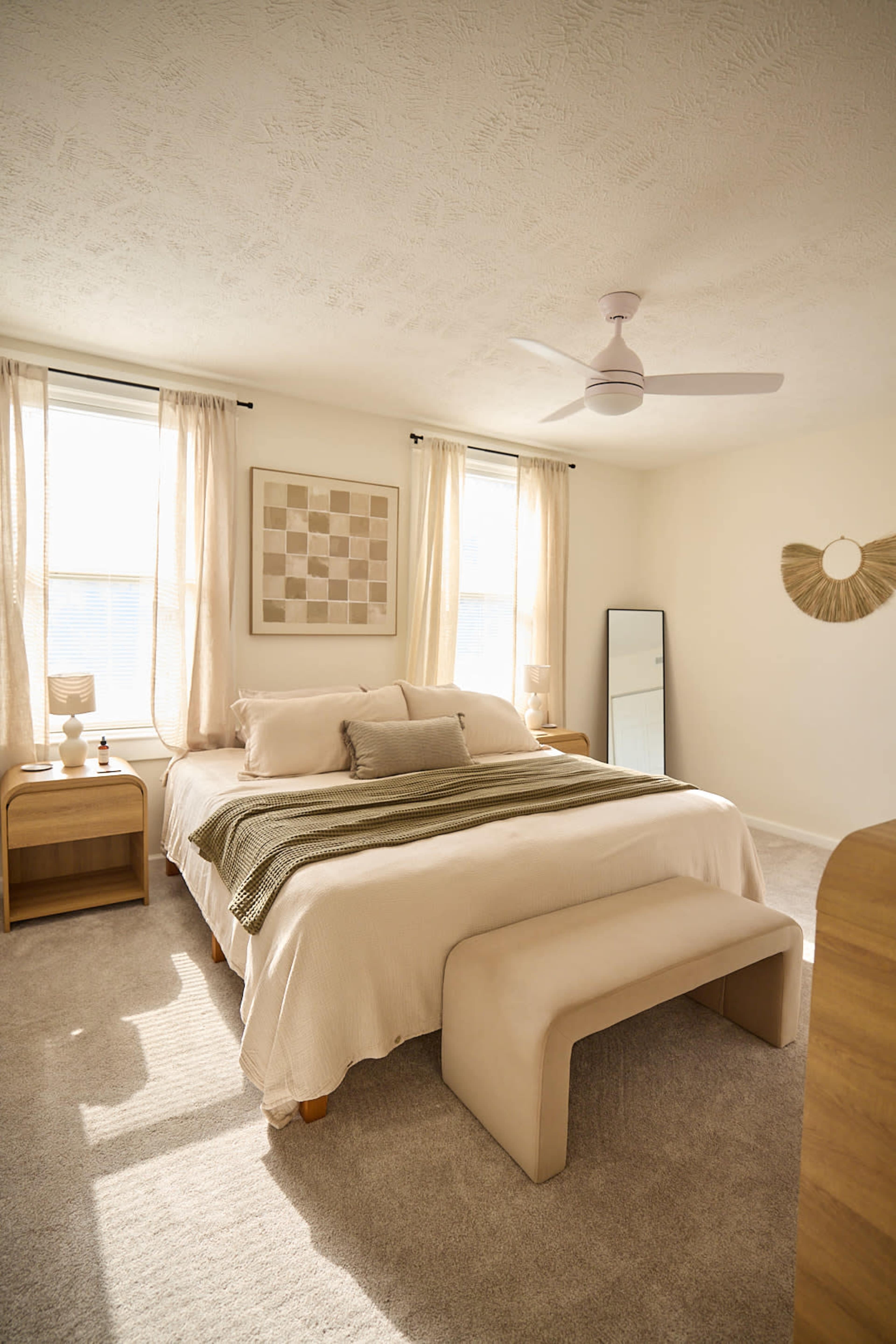 A well-lit bedroom features a large bed with neutral bedding, a bench at the foot, and minimalistic decor including a mirror and wall art.