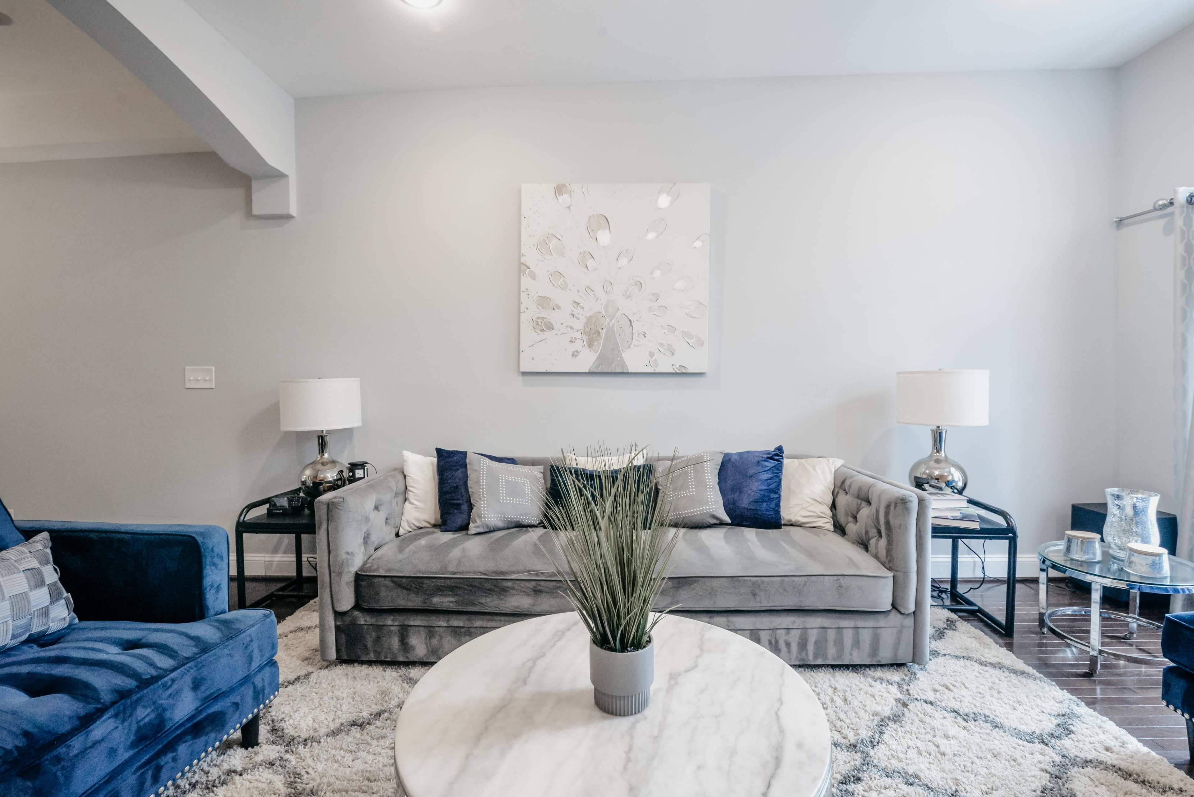 A modern living room features a gray sofa with decorative pillows, a round marble coffee table, and blue accent chairs, all highlighted by a wall art piece and two lamps.