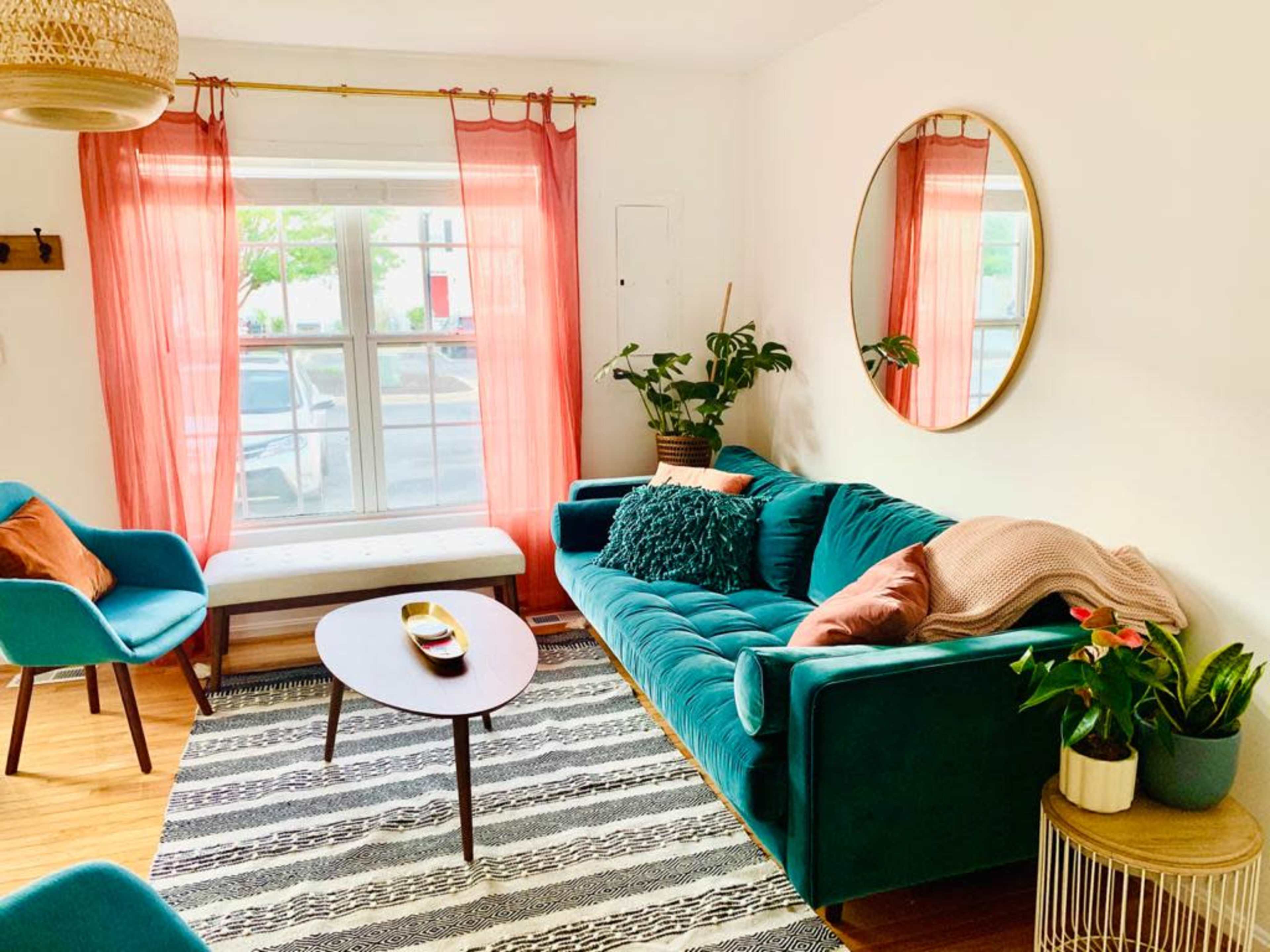 A cozy living room features a teal sofa, a round mirror, pink curtains, and a coffee table surrounded by plants and chairs.