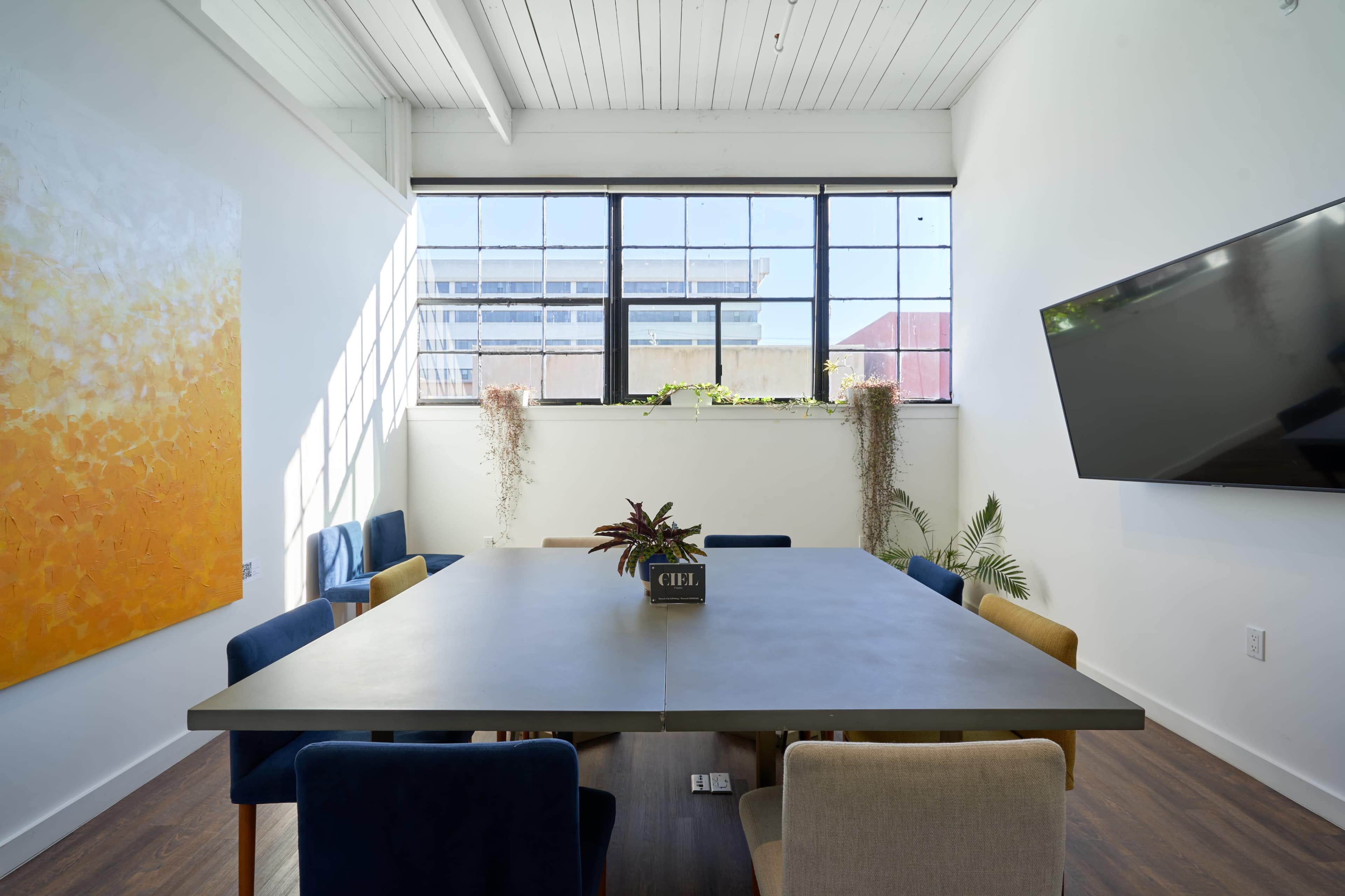 A conference room features a large table surrounded by several chairs, with a window allowing natural light and a colorful painting on the wall.