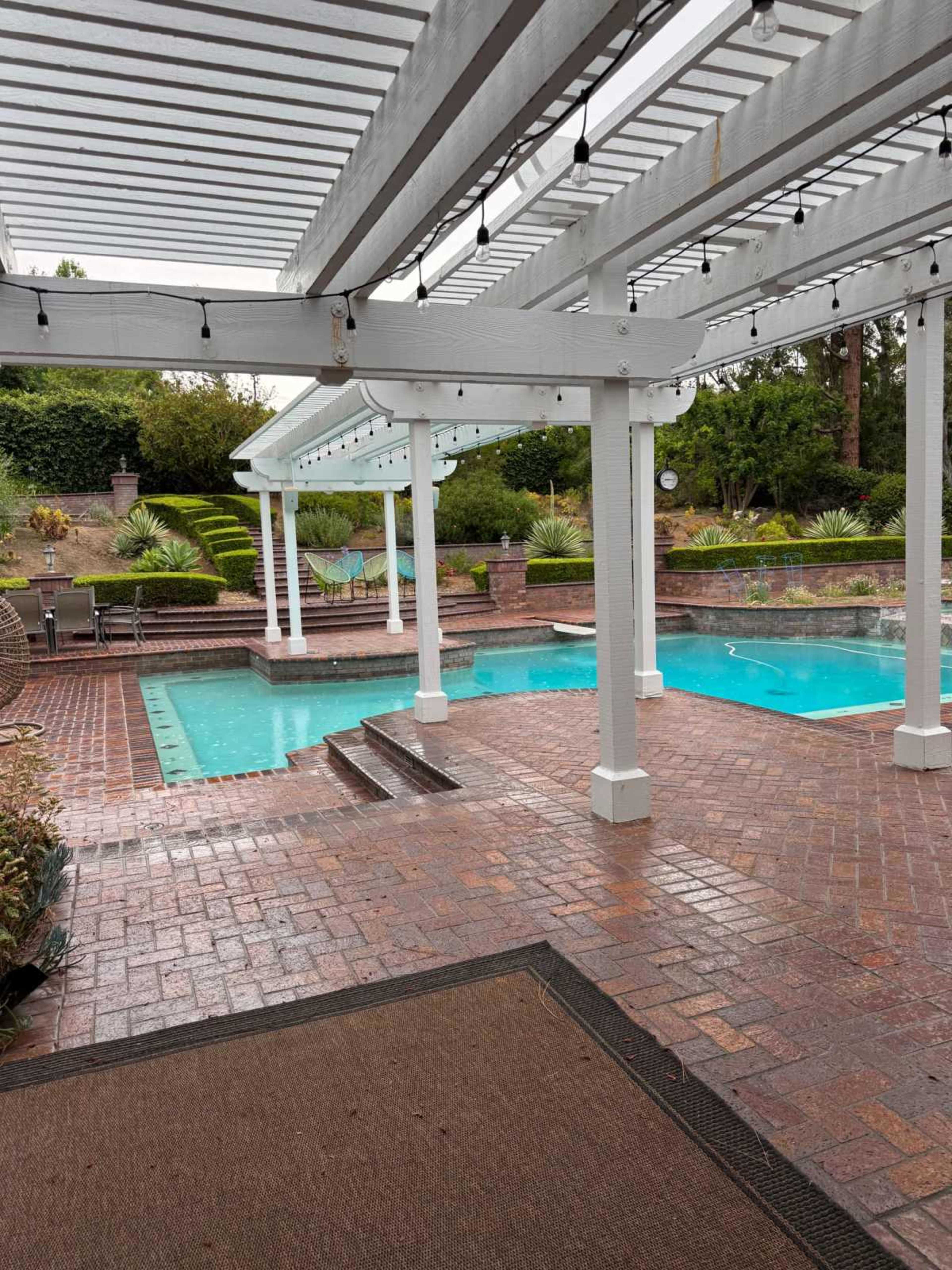 The image shows a covered patio area adjacent to a rectangular swimming pool, with a brick floor and landscaped greenery in the background.