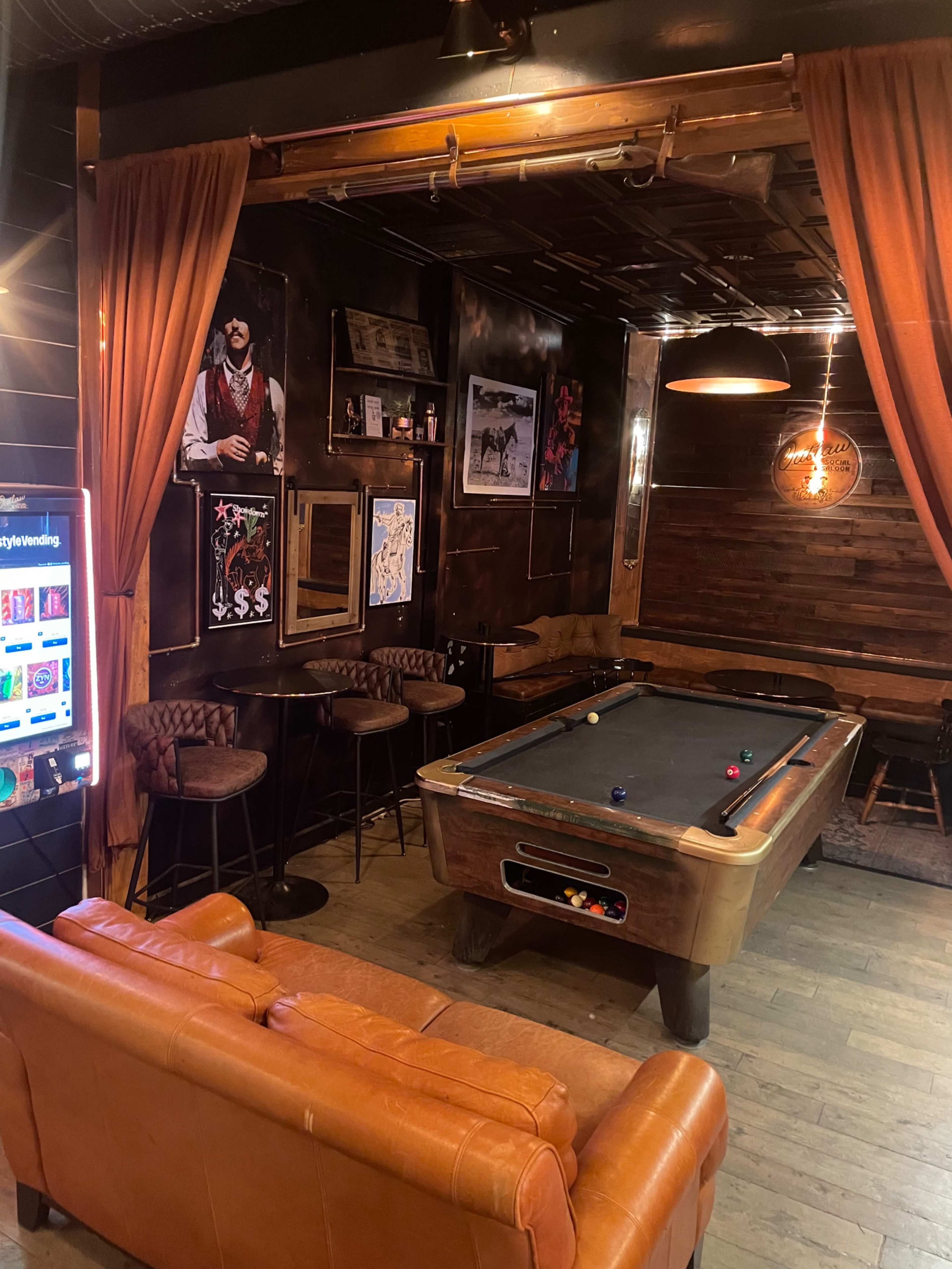 The image shows a cozy room featuring a billiards table surrounded by bar stools, a leather couch, and framed artwork on the walls.