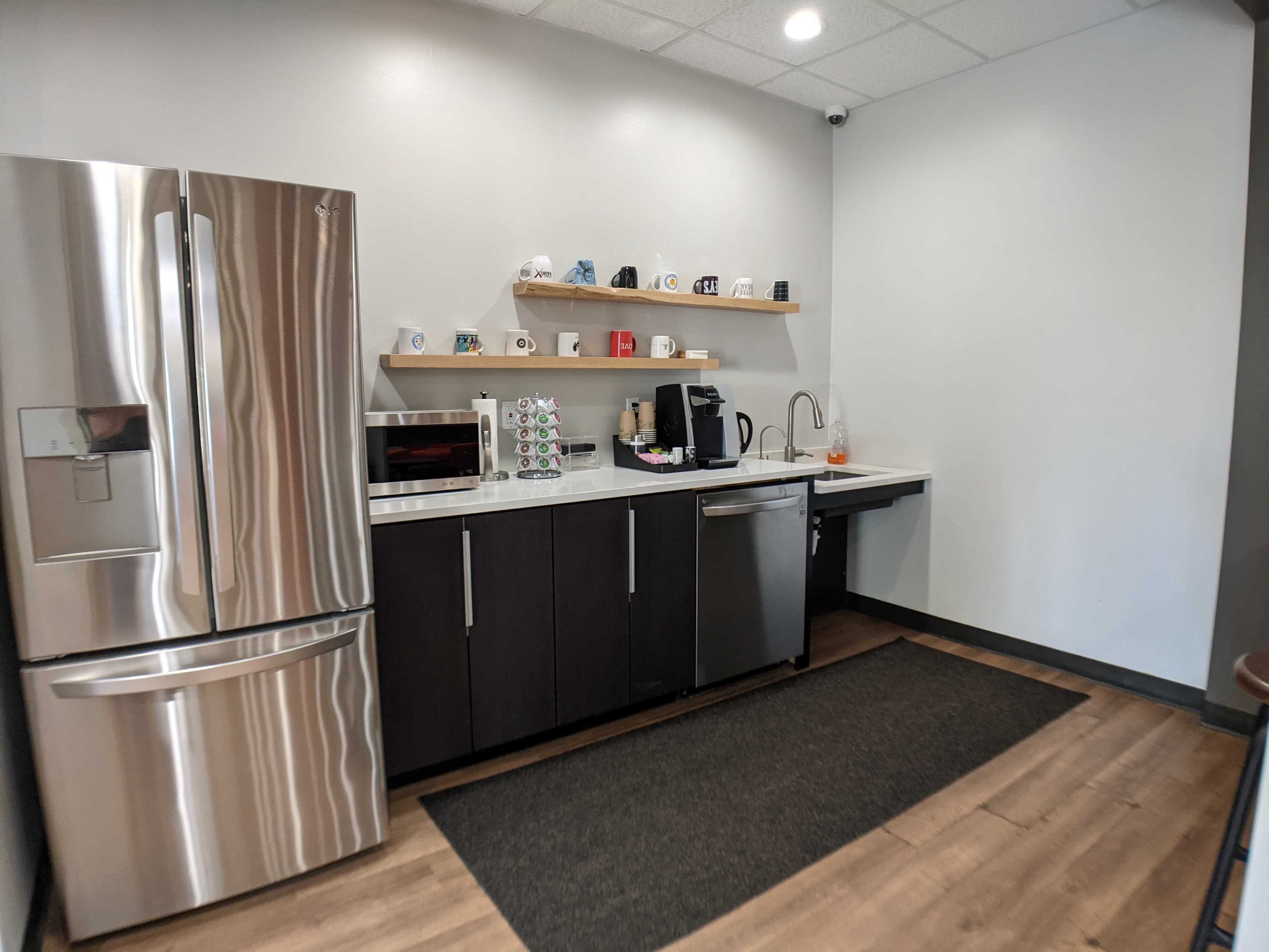 The image shows a modern kitchen area with a stainless steel refrigerator, a microwave, a coffee maker, and a sink, arranged along a dark cabinetry wall with shelves displaying mugs.