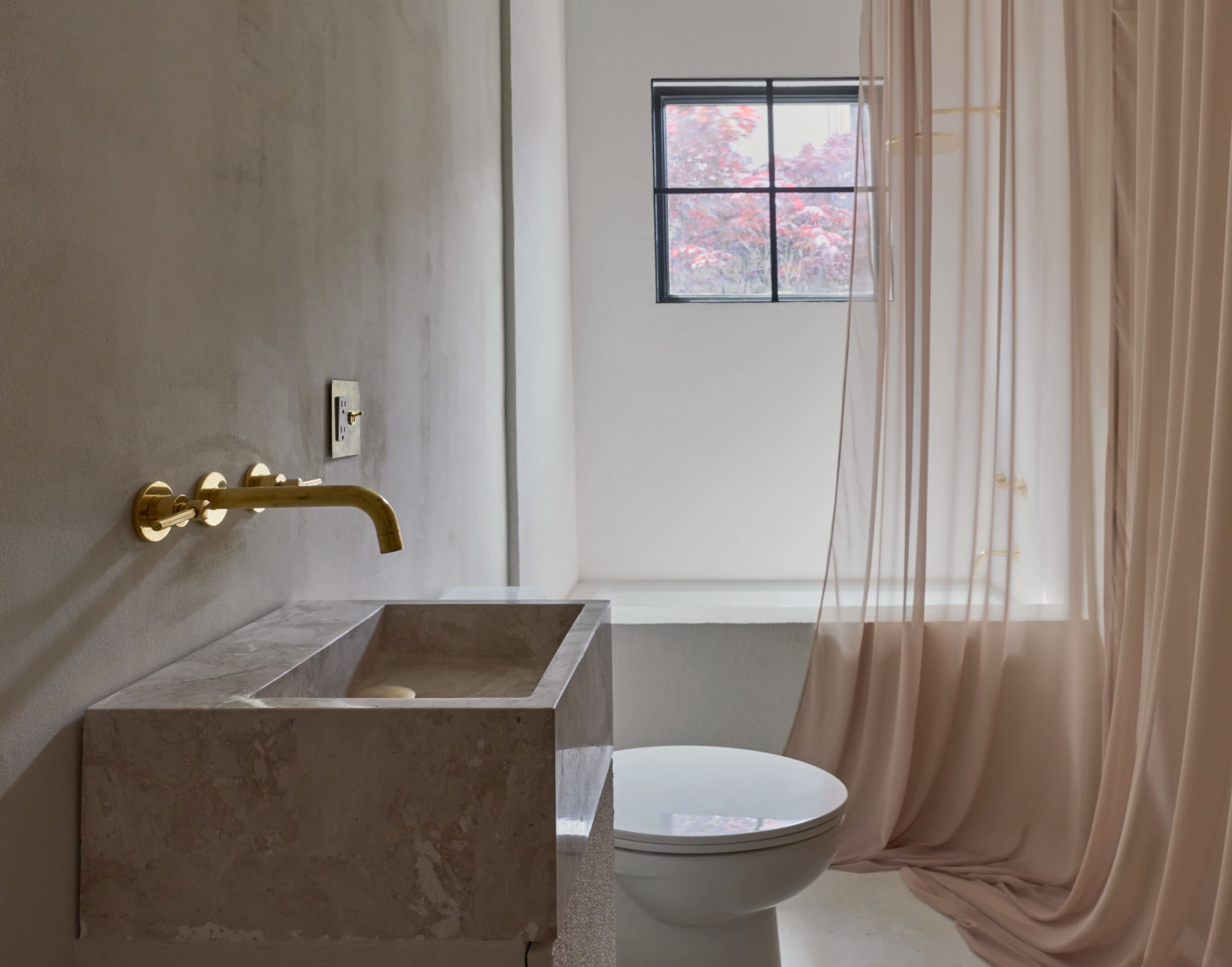 A modern bathroom featuring a gold faucet, a marble sink, a toilet, and a sheer curtain separating the space from a window with a view of red foliage outside.