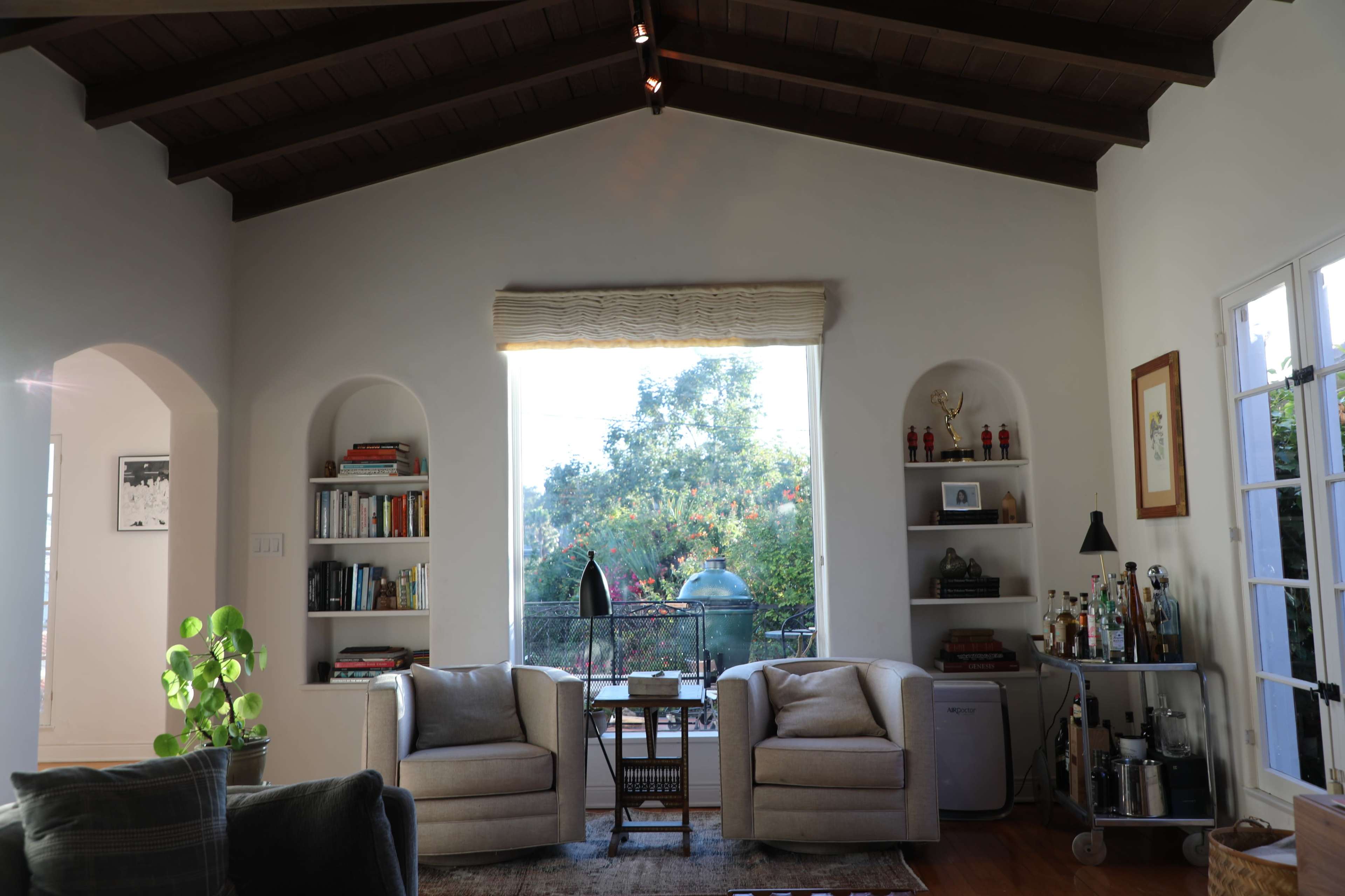 A cozy living room features two armchairs facing a large window with a view of greenery, complemented by shelves of books and decorative items.