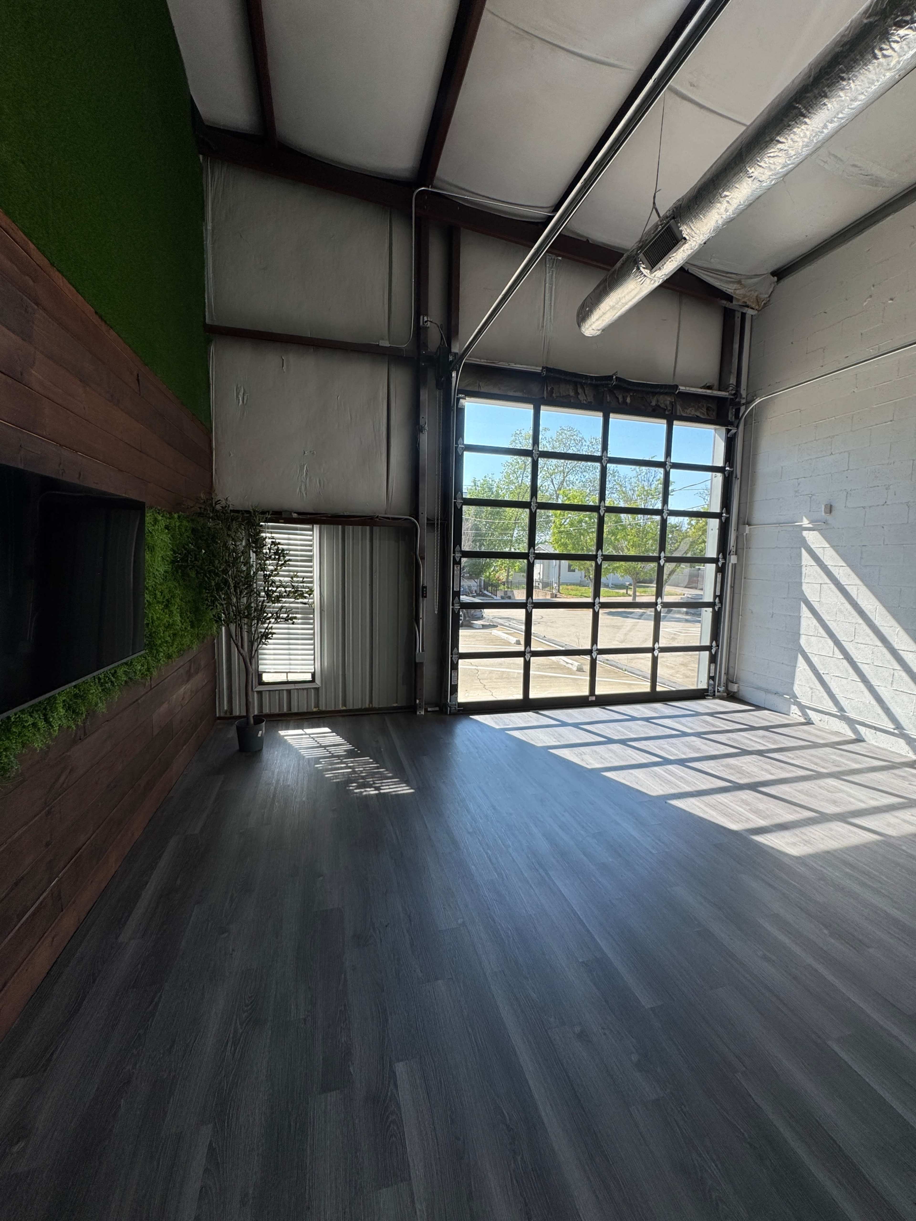 The image shows a spacious, well-lit interior of a building with a large glass garage door, wooden paneling, and a patch of artificial grass on the wall.