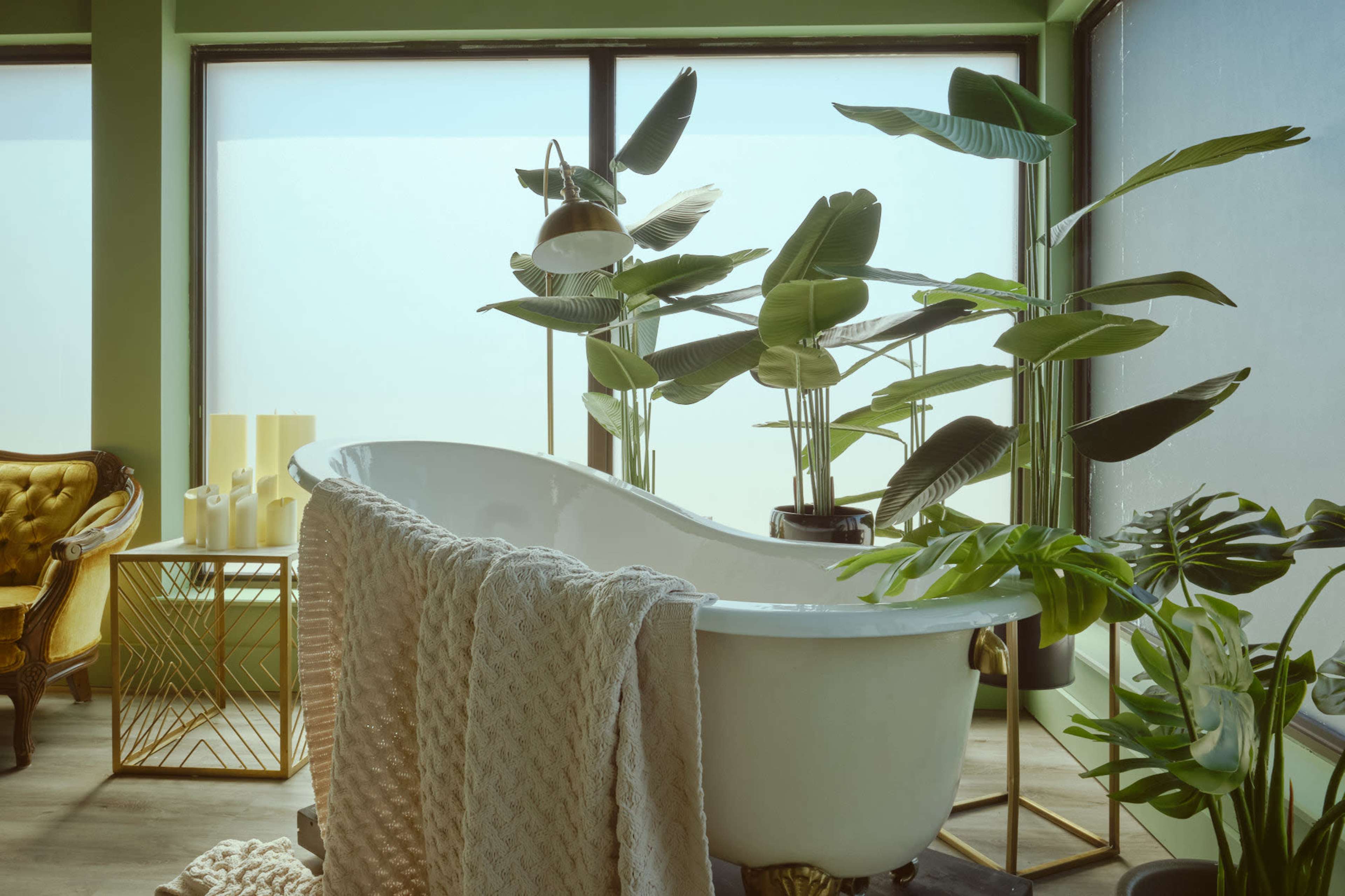 The image features a stylish bathroom with a white freestanding bathtub surrounded by potted plants and a decorative armchair.