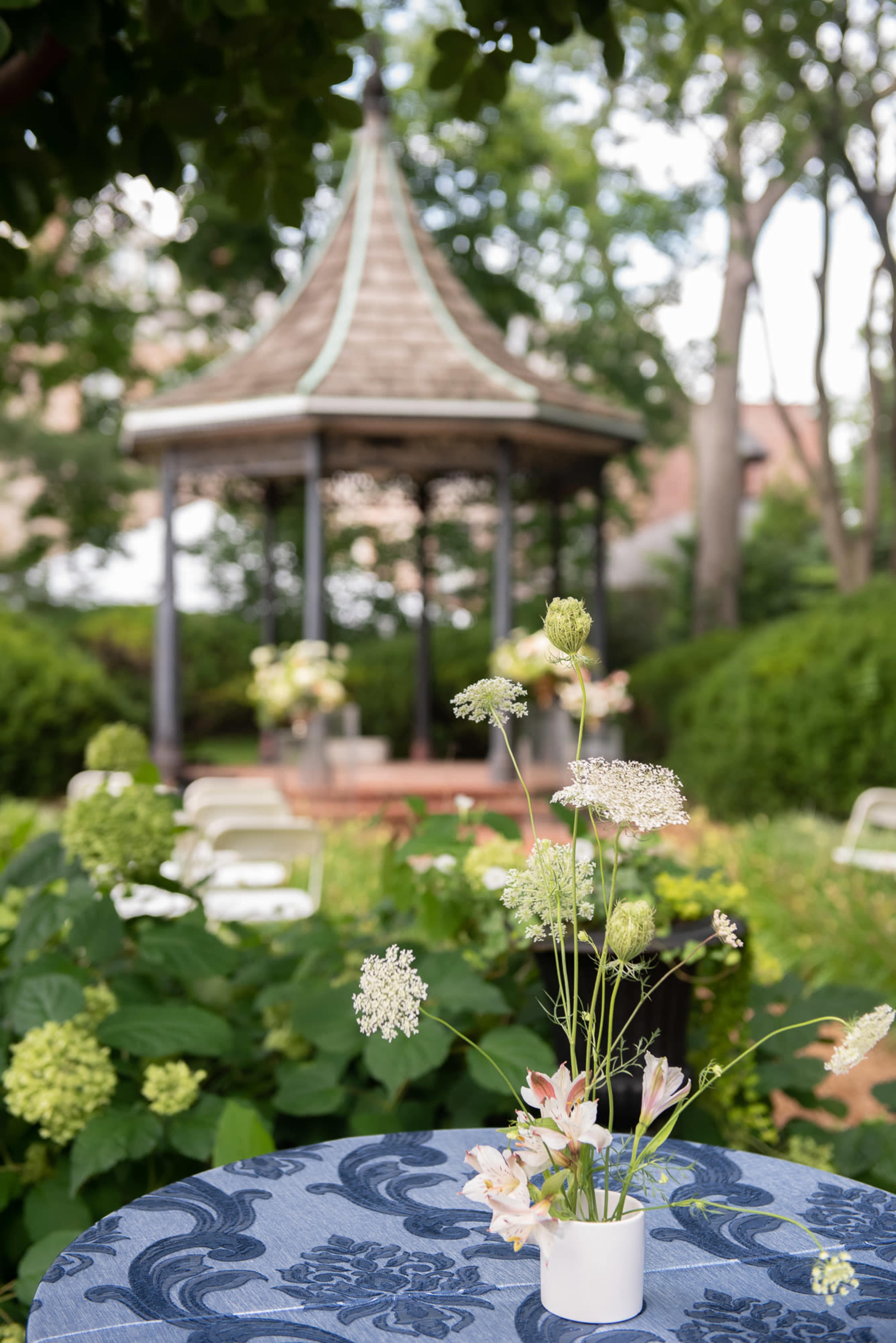A table with a floral centerpiece is set in a garden featuring a gazebo in the background.