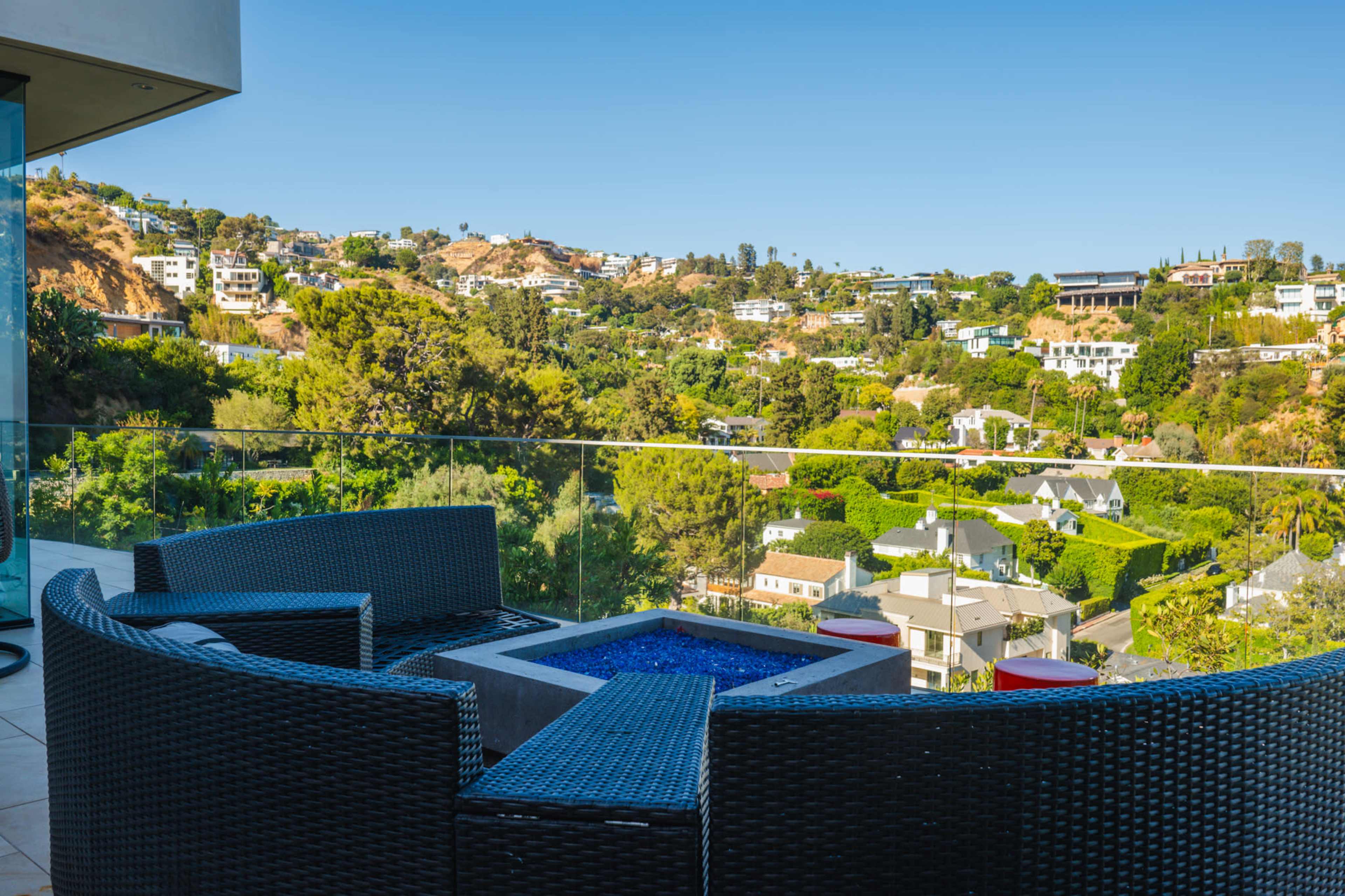 The image shows a balcony with a circular seating area and a small fire pit, overlooking a hillside dotted with residential homes and trees.