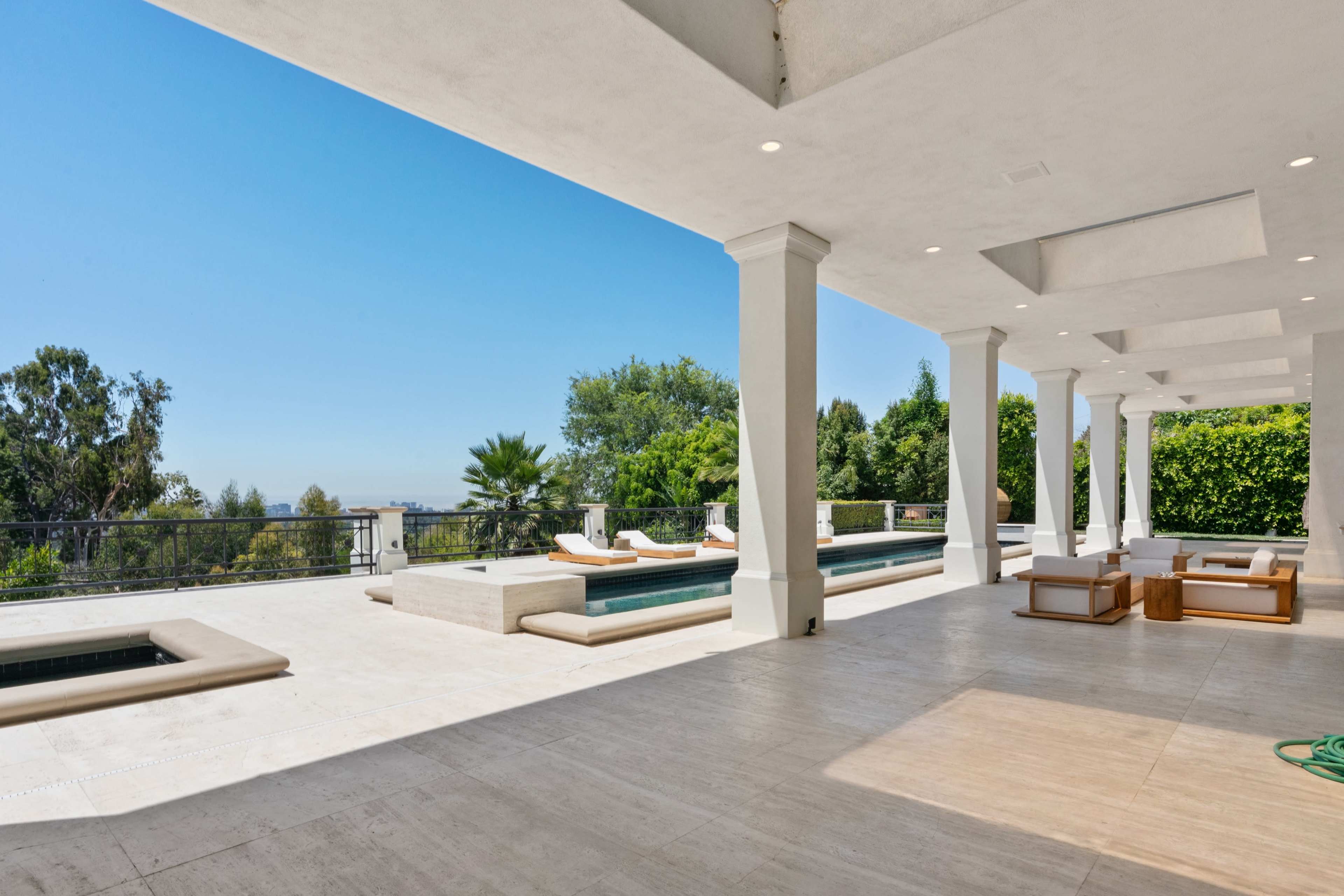 A spacious outdoor patio area featuring a pool and sun loungers, surrounded by greenery under a clear blue sky.