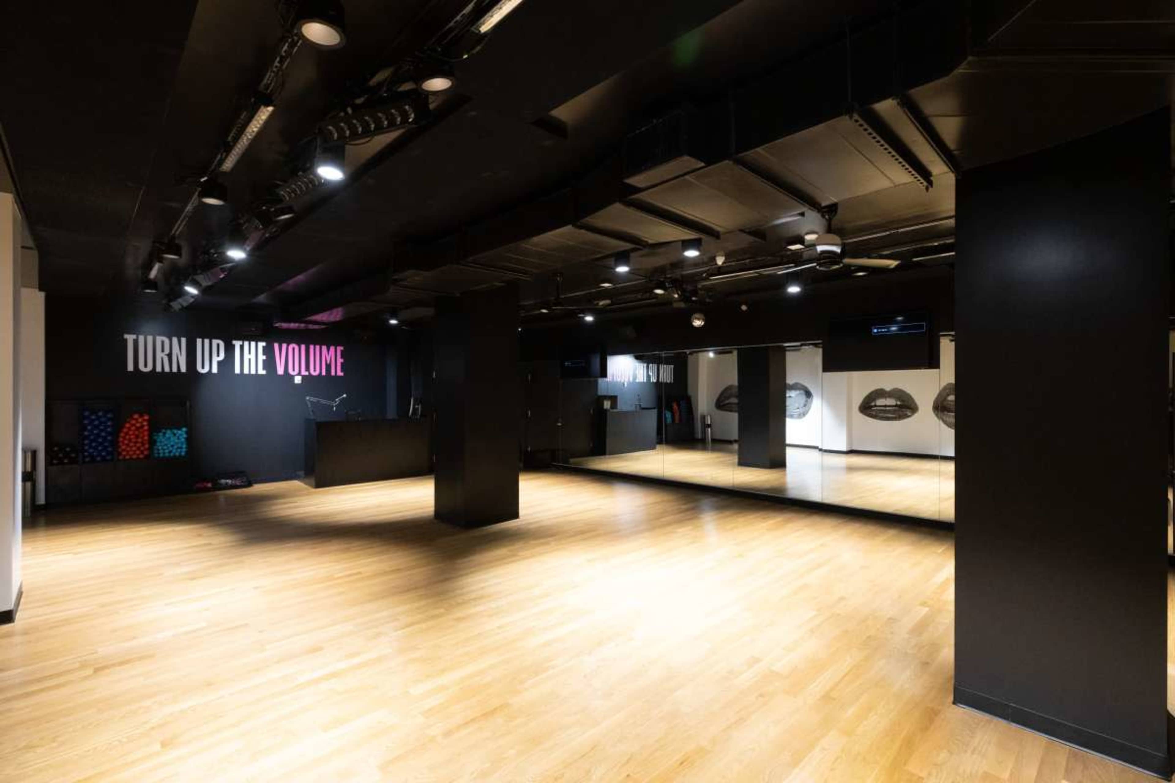 The image shows a spacious dance studio with wooden flooring, black walls, and a large mirror on one side.