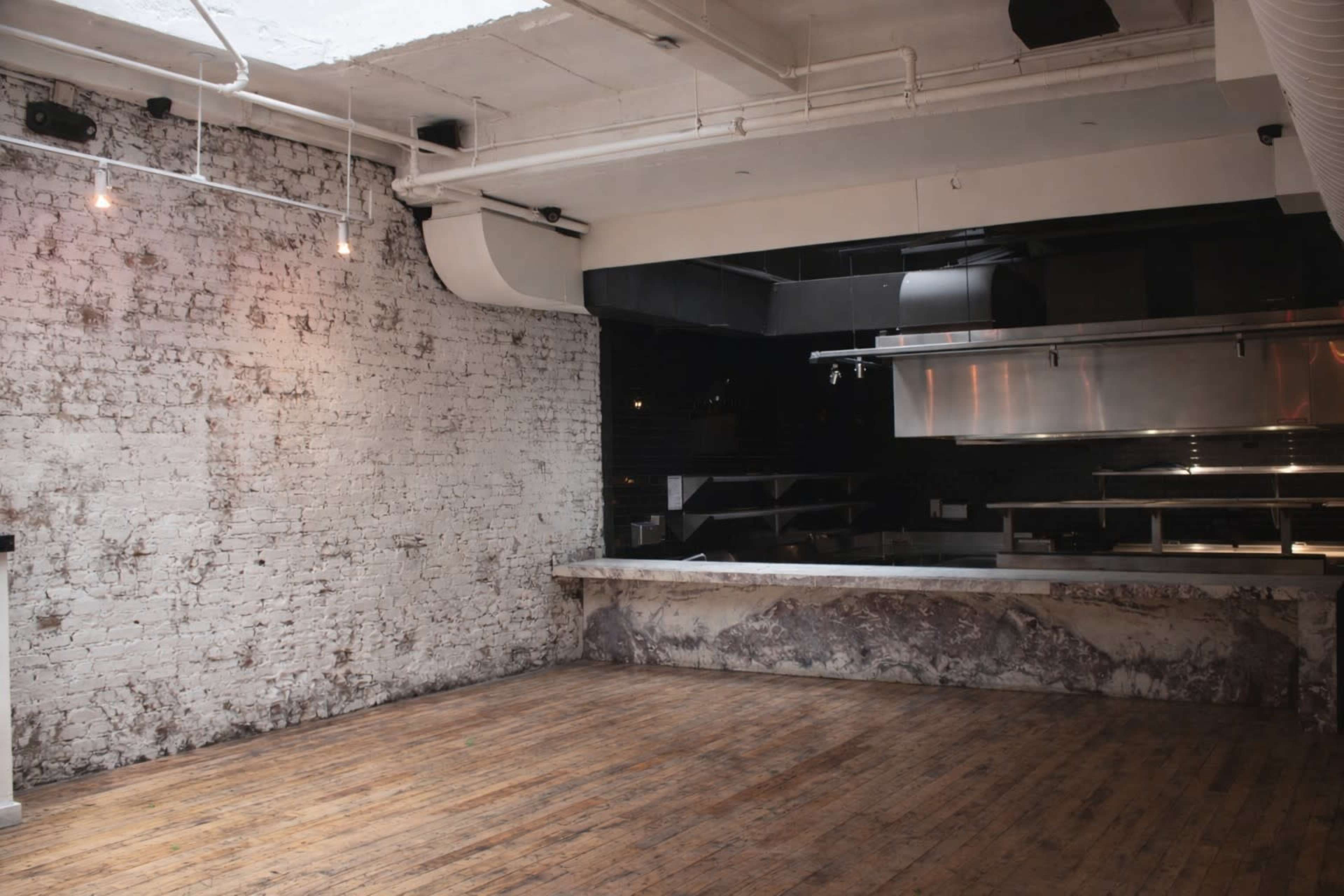 The image shows an empty interior space with exposed brick walls, wooden flooring, and kitchen equipment in the background.