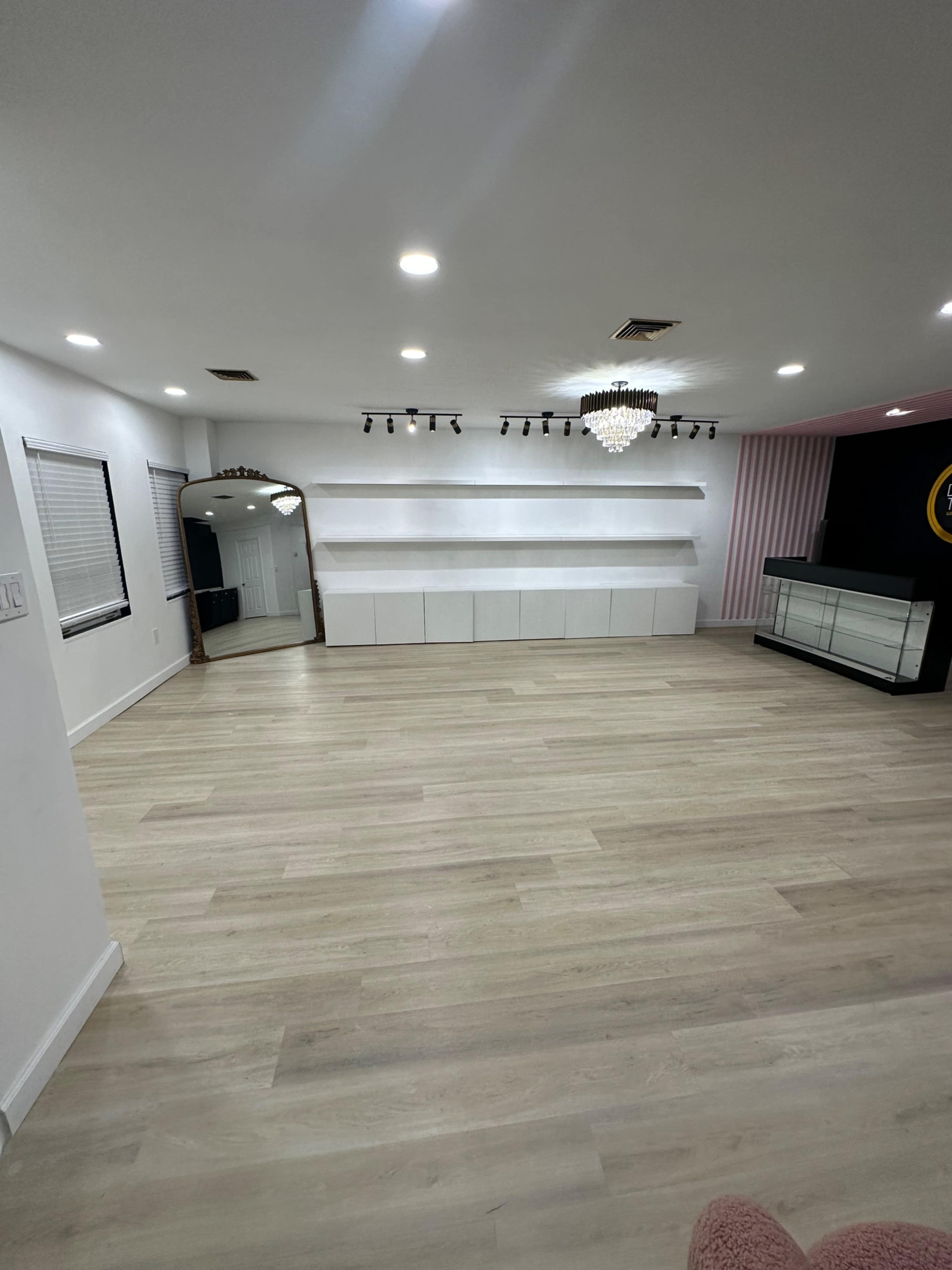 The image shows a spacious, well-lit room with light wood flooring, featuring a large mirror on one side and a white shelving unit against the back wall.