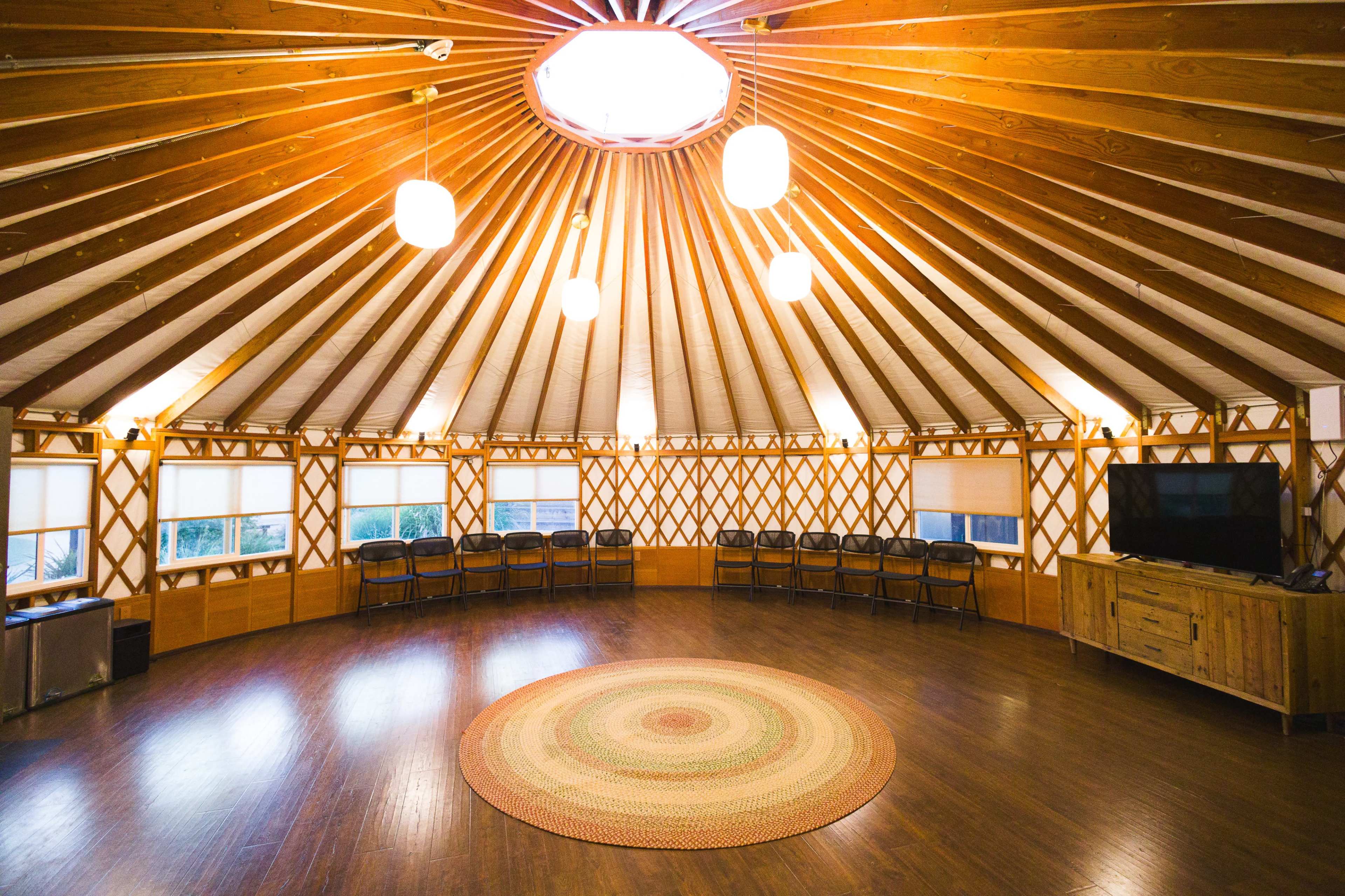 The interior of a round yurt features wooden beams, circular skylight, and a central area with a circular rug surrounded by chairs.