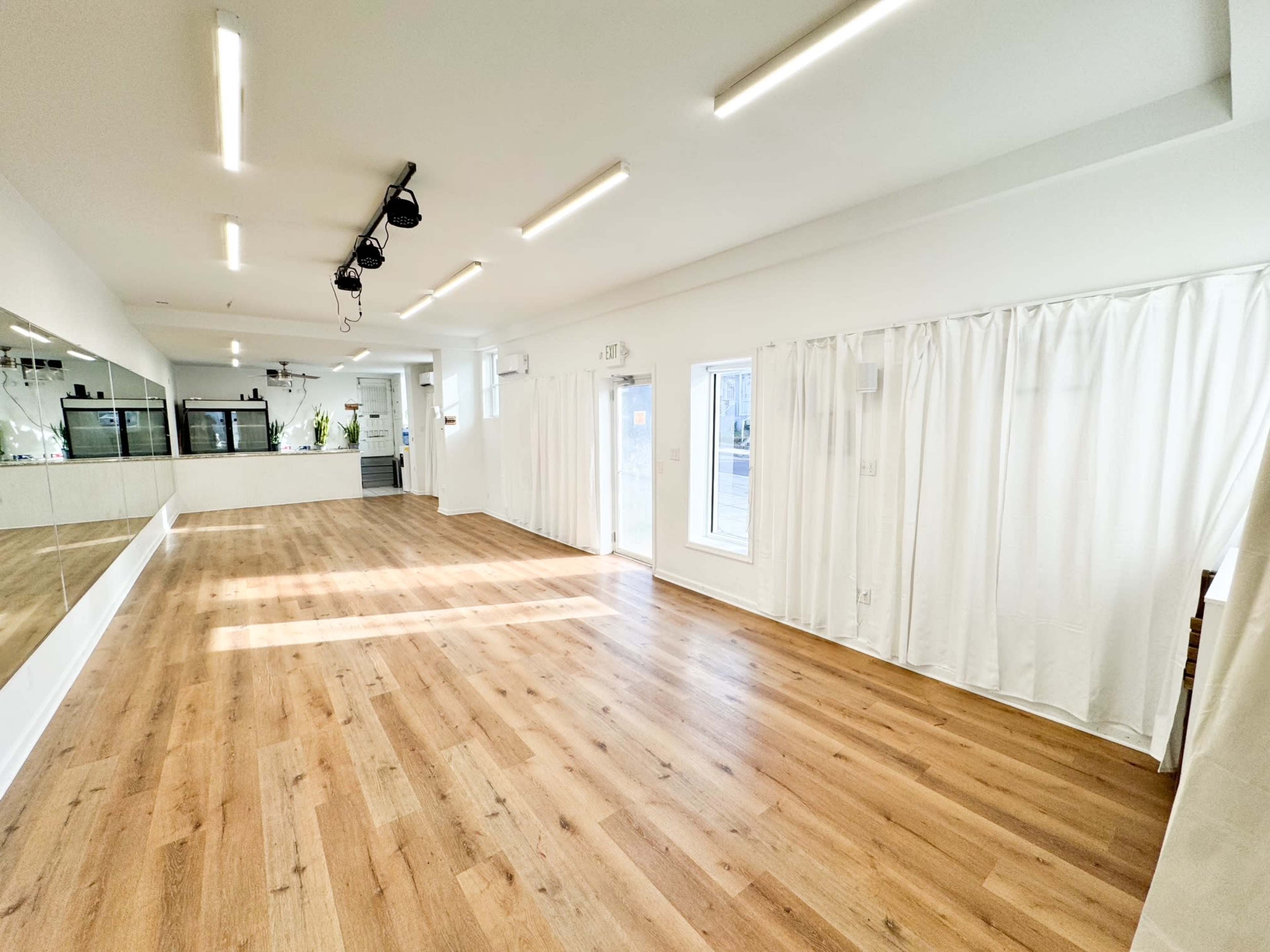 The image shows a spacious room with wooden flooring, white walls, and large windows, featuring a mirror along one side and sheer curtains.