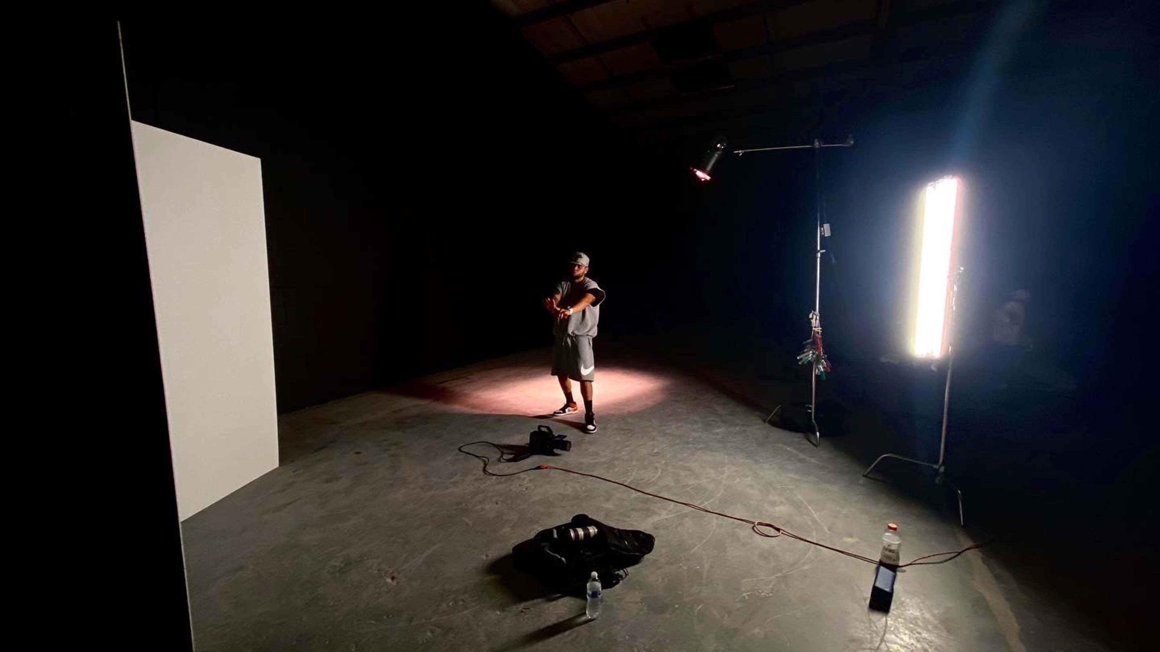 A person stands in a dimly lit studio with a violin, near a white panel and some lighting equipment.