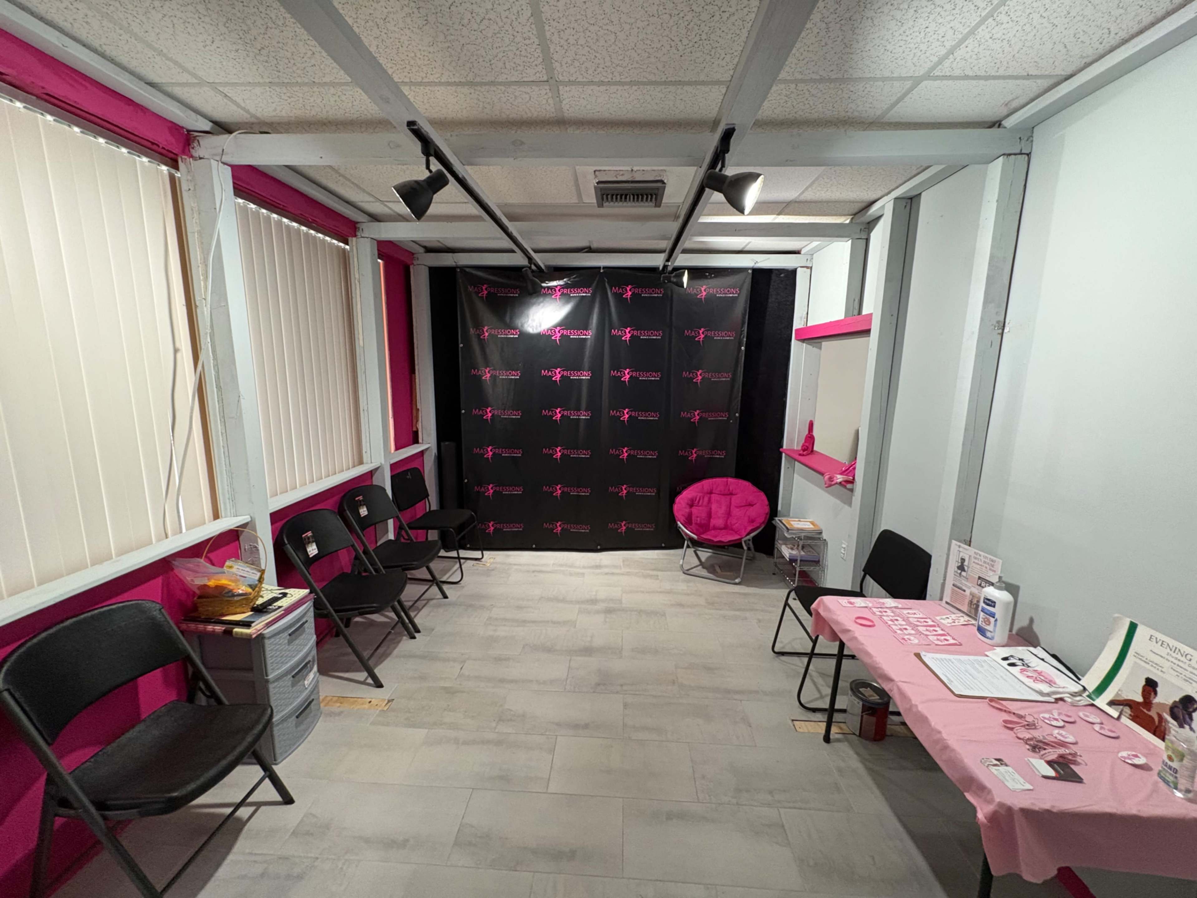 The image shows a room with pink accents, featuring several black chairs arranged facing a dark backdrop, a round pink chair, and a table covered with a pink cloth displaying pamphlets and materials.