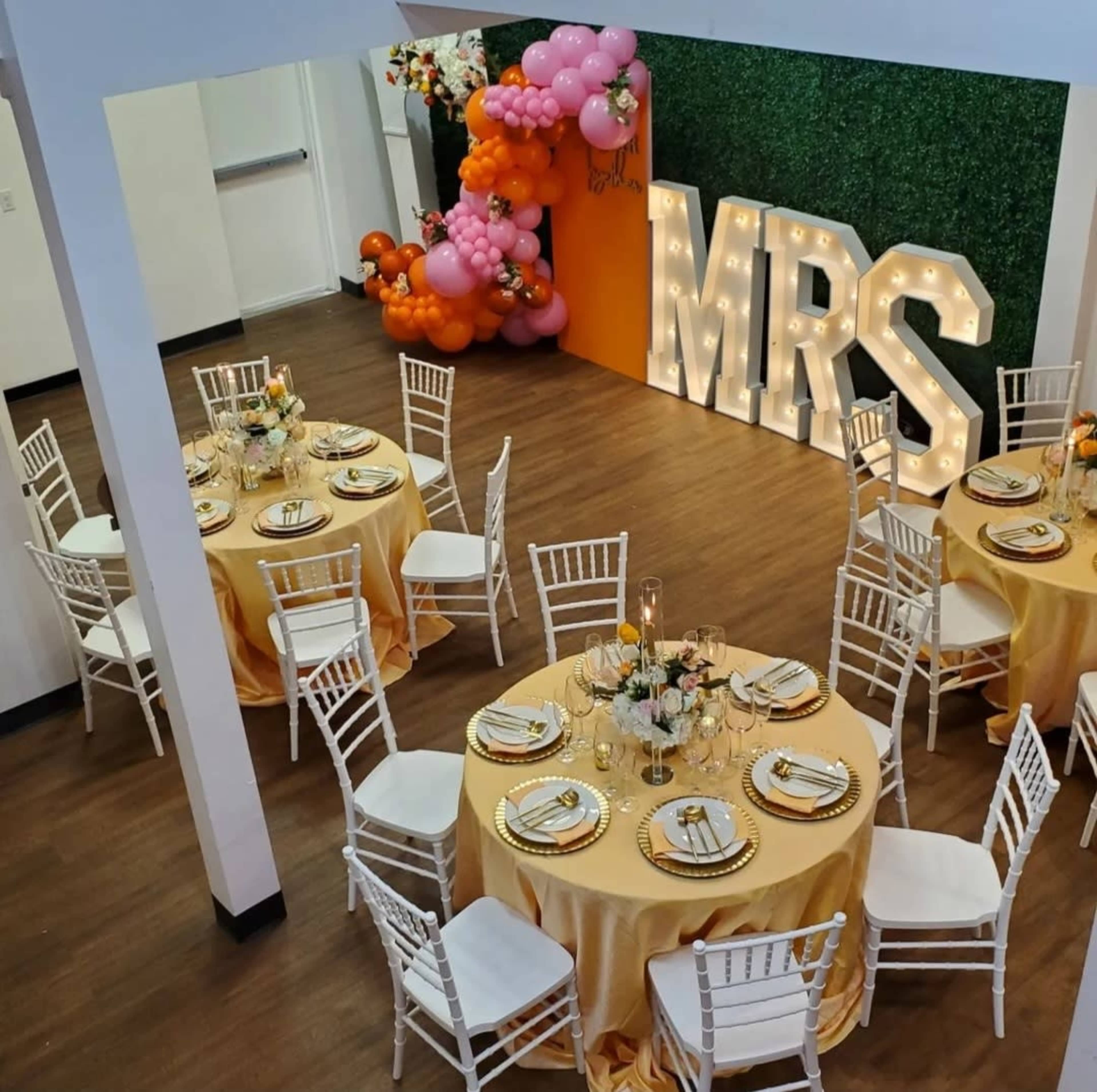 A decorated event space with round tables covered in yellow tablecloths and a large floral backdrop featuring the letters "MRS."