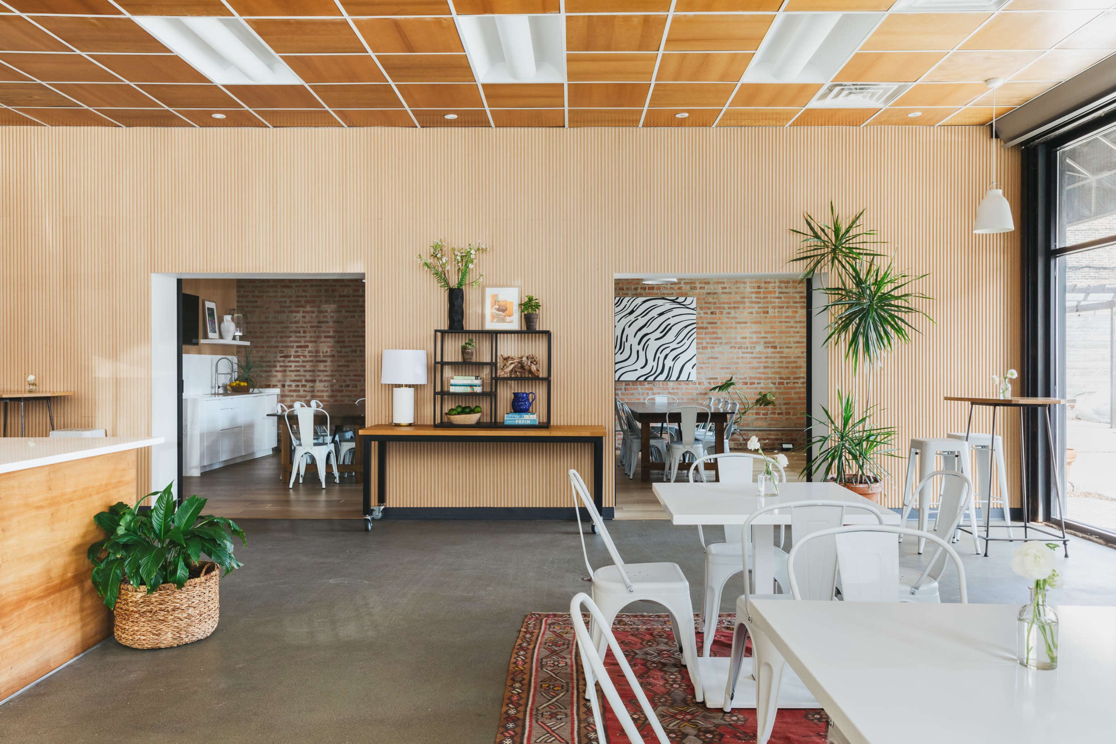 The image shows a modern café with wooden paneling, white tables and chairs, indoor plants, and a patterned wall art.