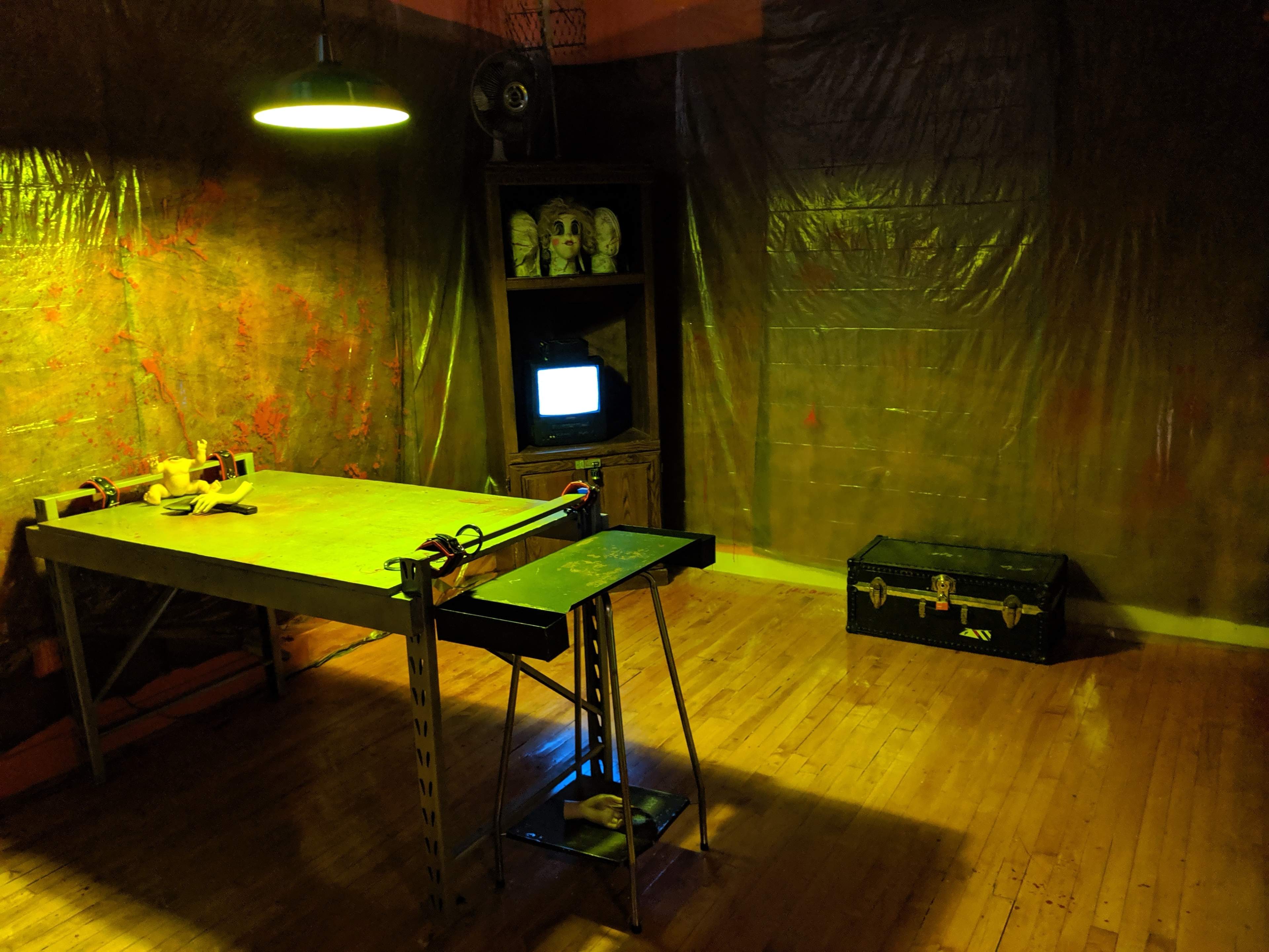 The dimly lit room features a table with tools, a vintage television set, and a dark trunk, all against plastic-covered walls.
