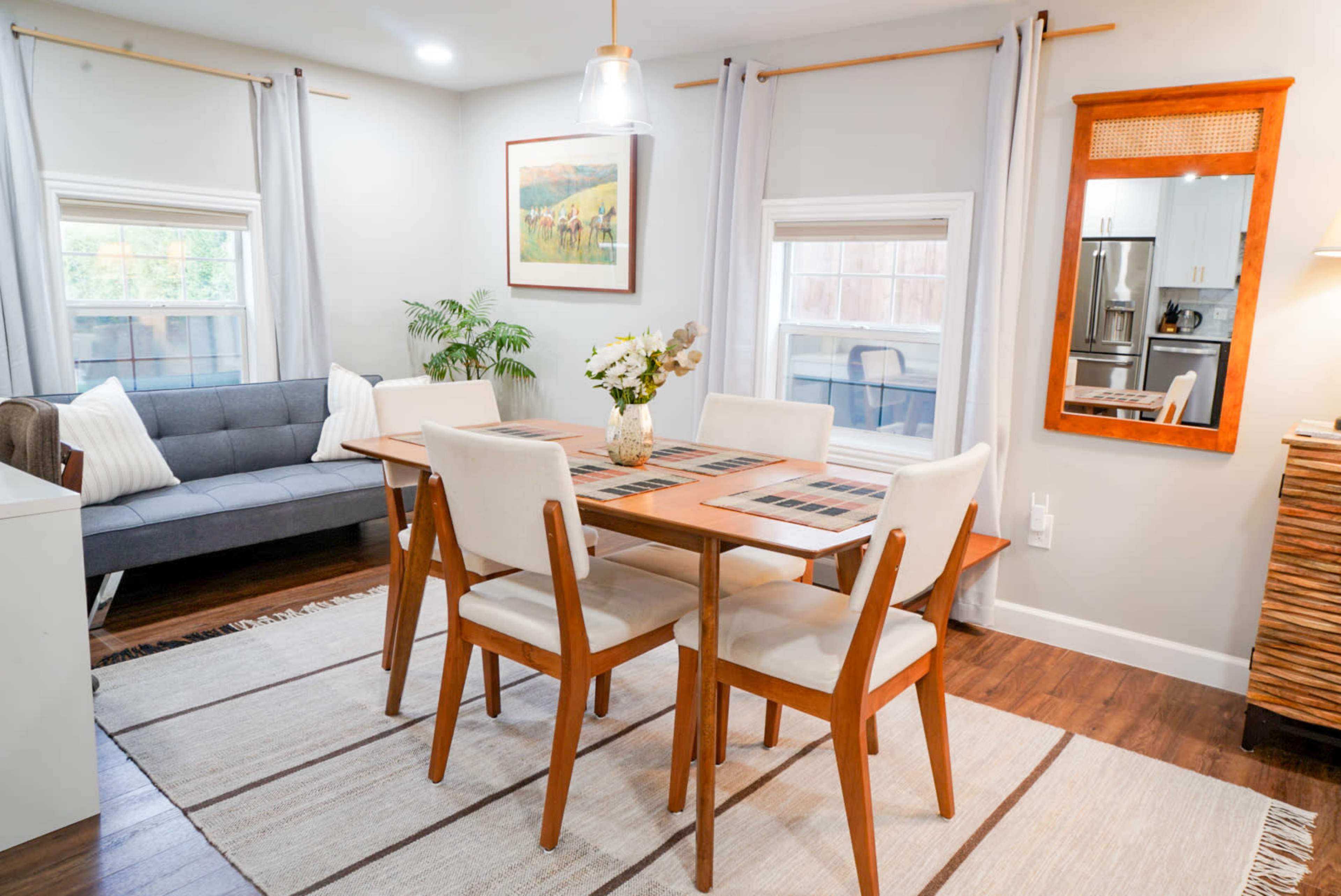 A dining area features a wooden table surrounded by four chairs, a gray sofa in the corner, and large windows with light curtains.