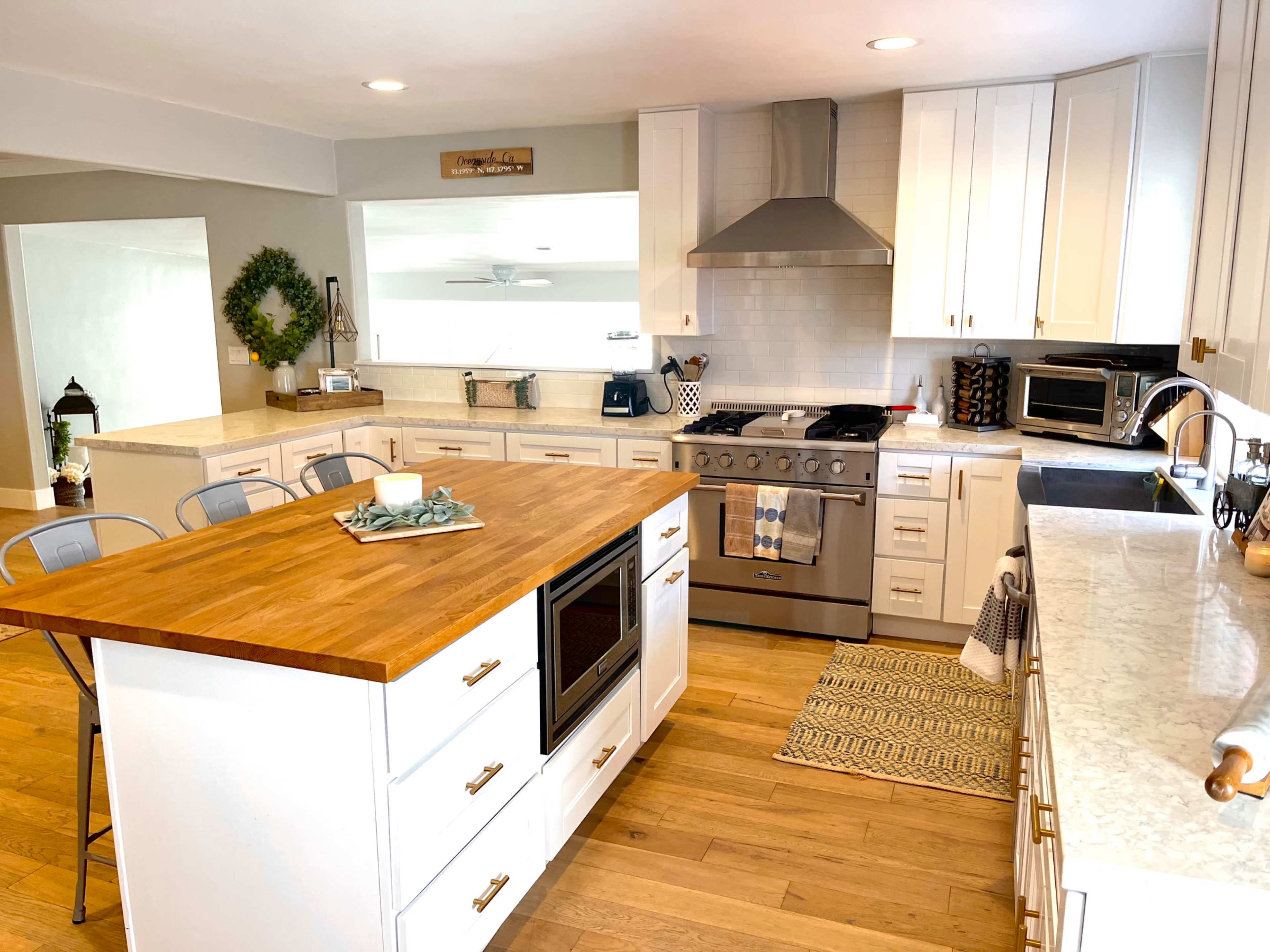 A modern kitchen with a wooden island, stainless steel appliances, and white cabinetry, illuminated by natural light from a nearby window.