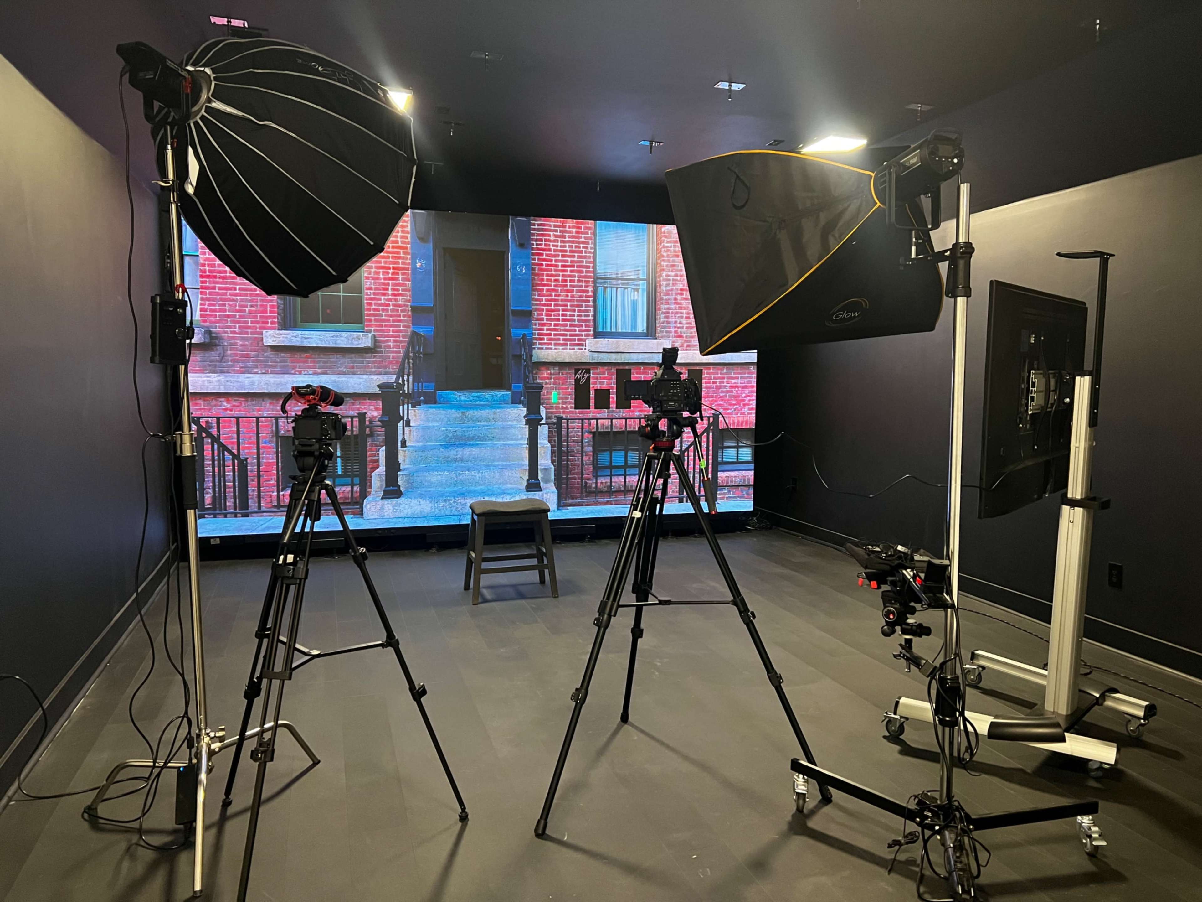 A studio setup with two tripods holding cameras, various lighting equipment, and a large screen displaying a brick building.