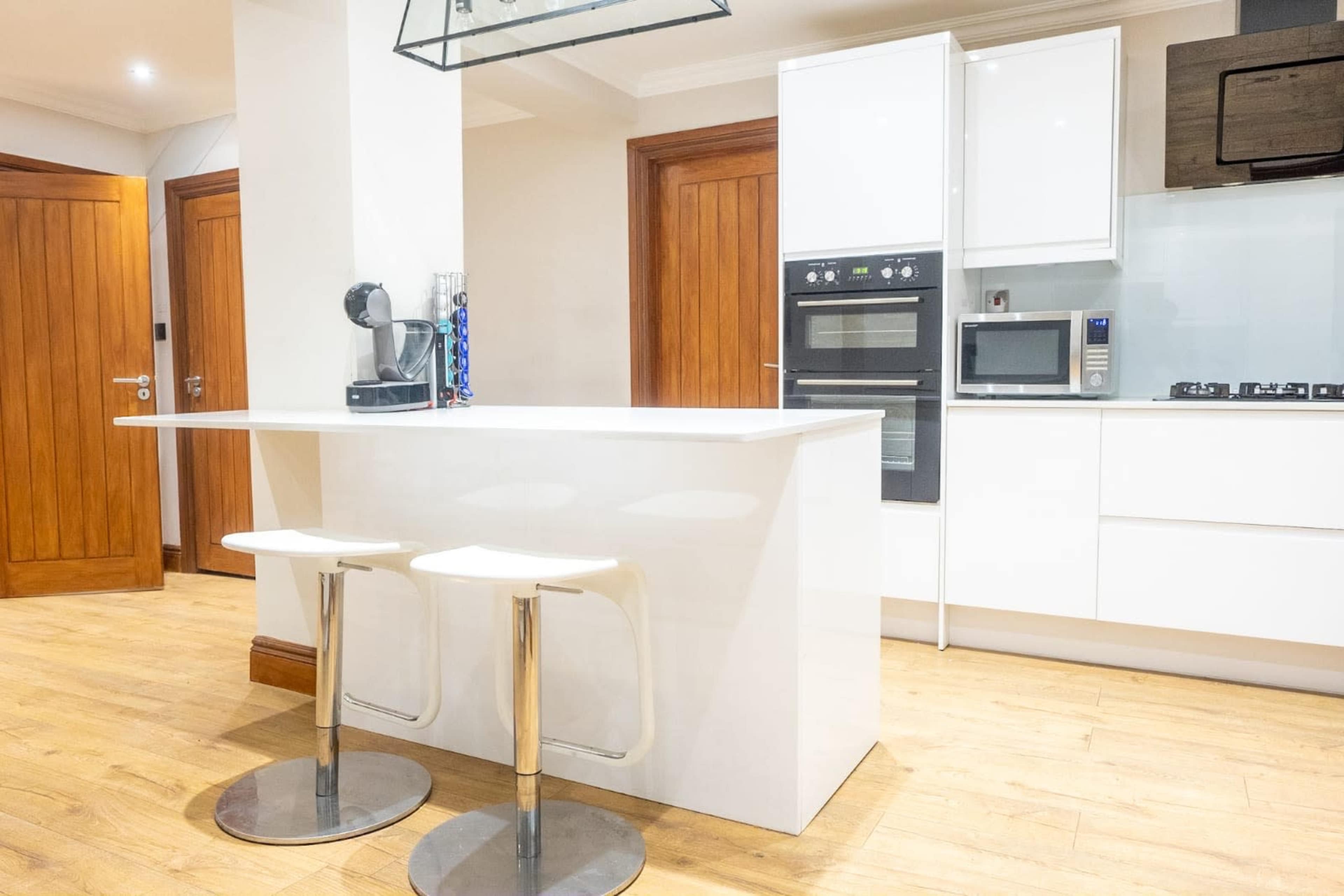 The image shows a modern kitchen with a white bar counter and two stools, equipped with built-in appliances and wooden cabinetry.