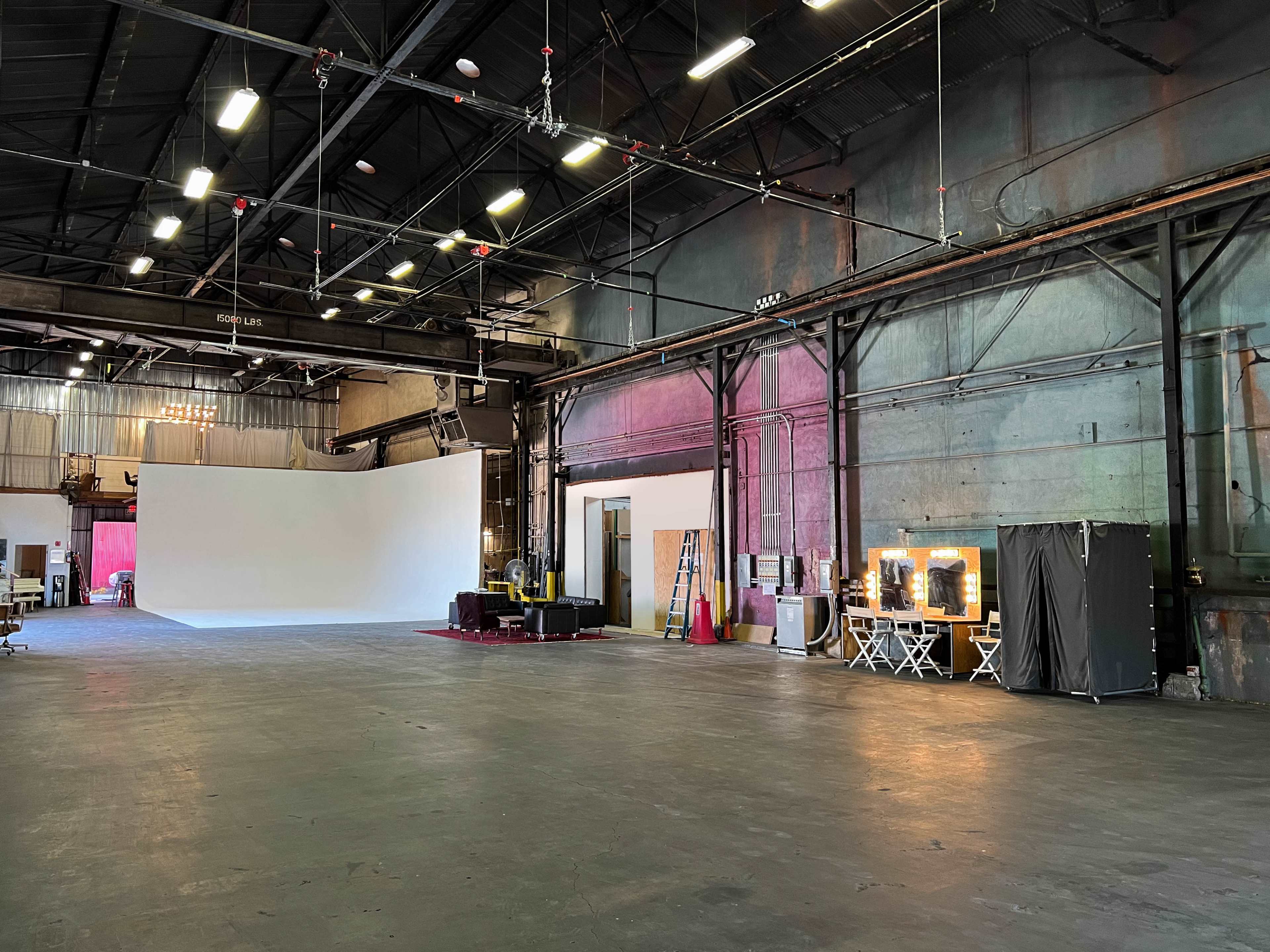 The image shows a spacious industrial warehouse with a large white backdrop, a makeup station with mirrors, and various equipment along one side.
