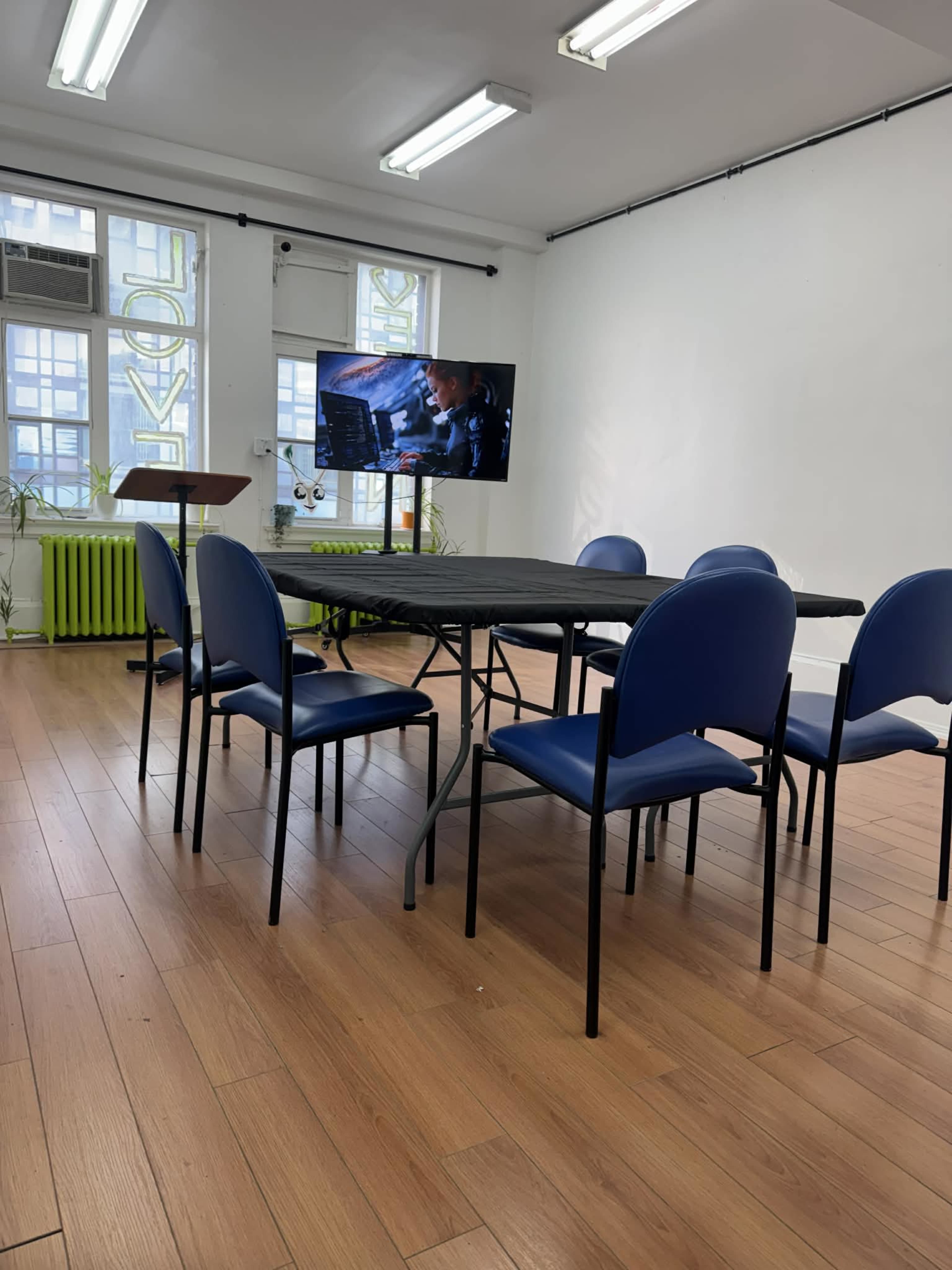 The image shows an empty meeting room with a rectangular table surrounded by blue chairs and a television displaying a scene on one wall.