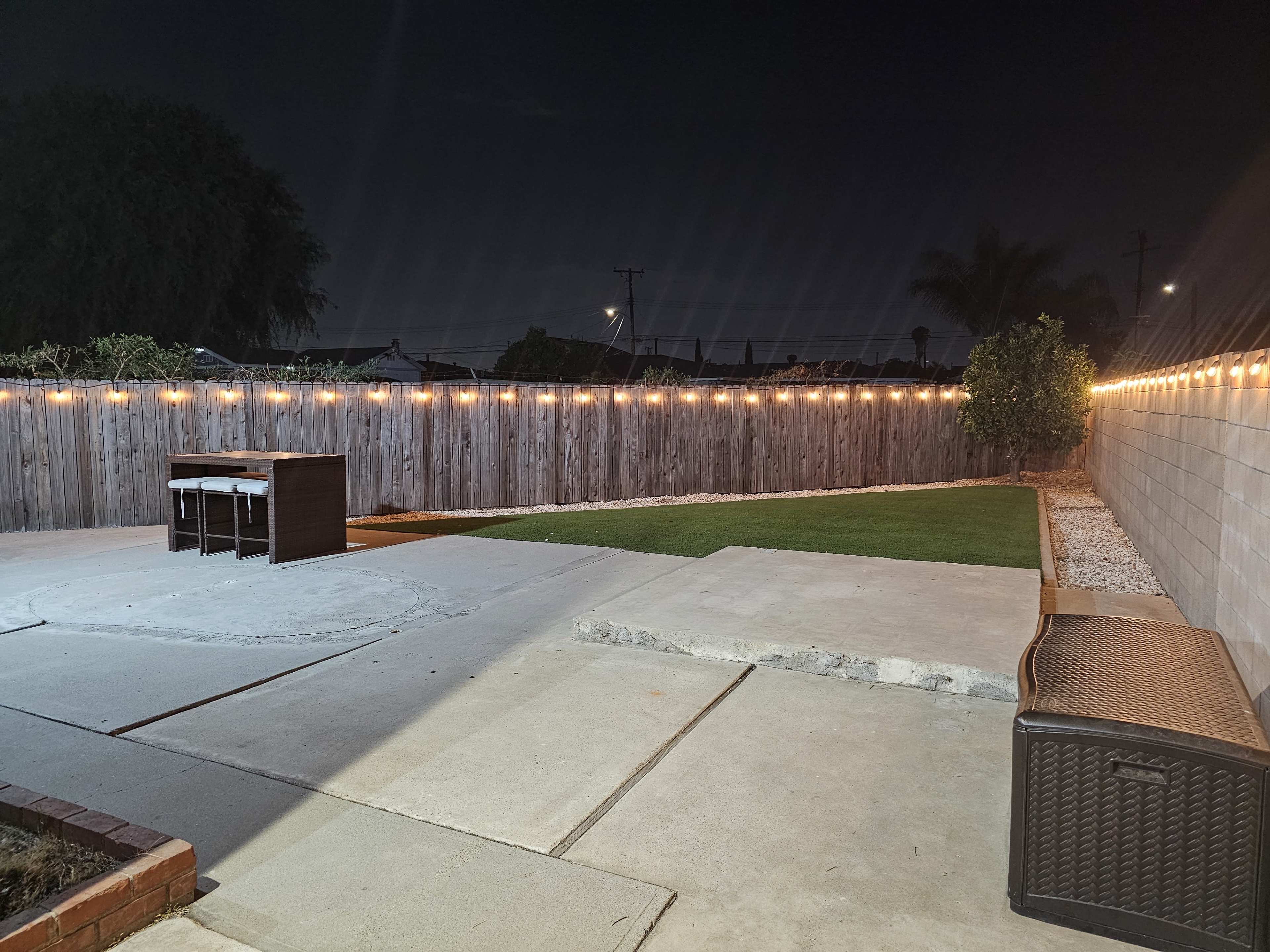 A backyard at night with string lights illuminating a concrete patio area and a small table set against a wooden fence.