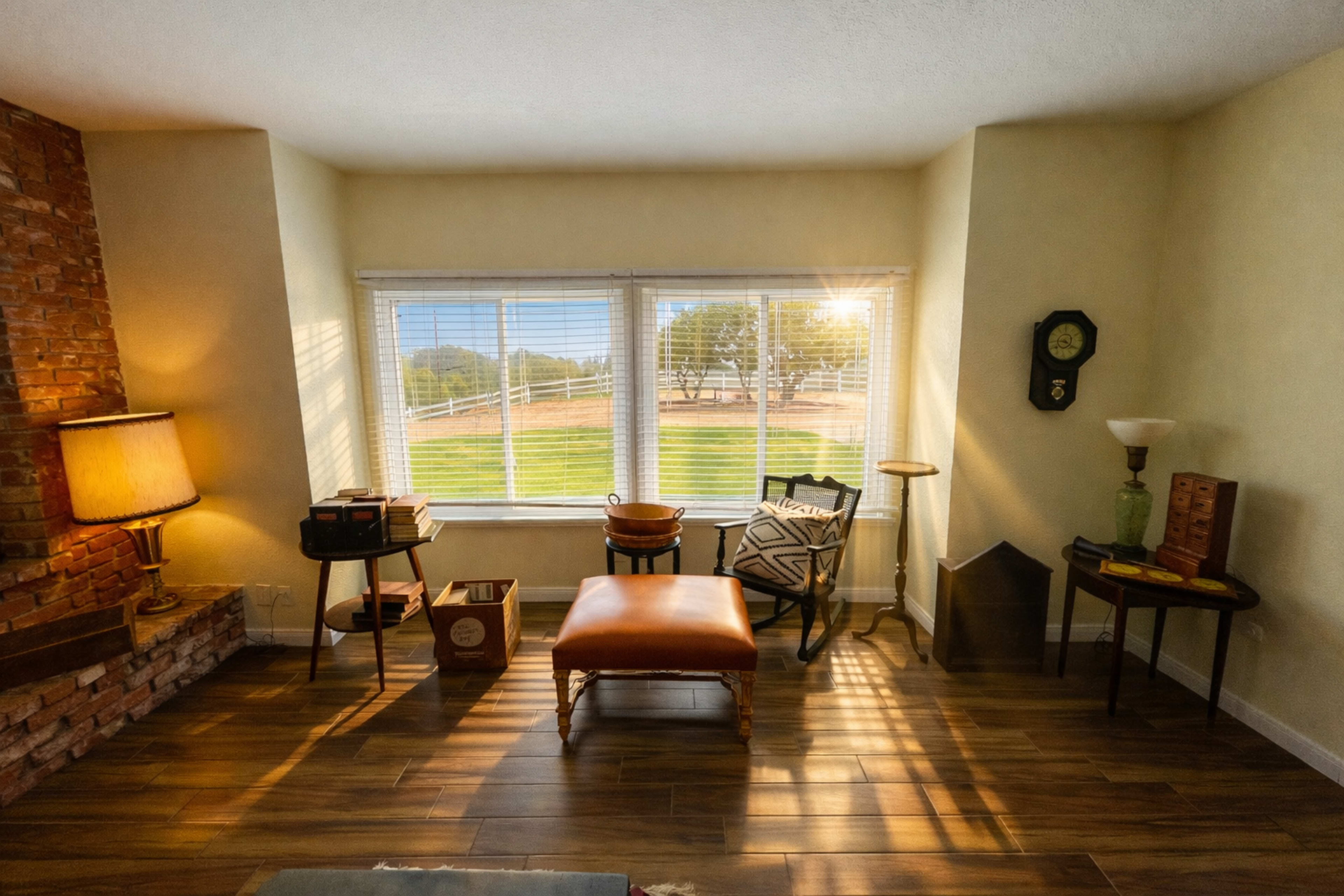 A cozy room features a brick wall, a lamp, various pieces of furniture, and large windows that overlook a grassy outdoor area.