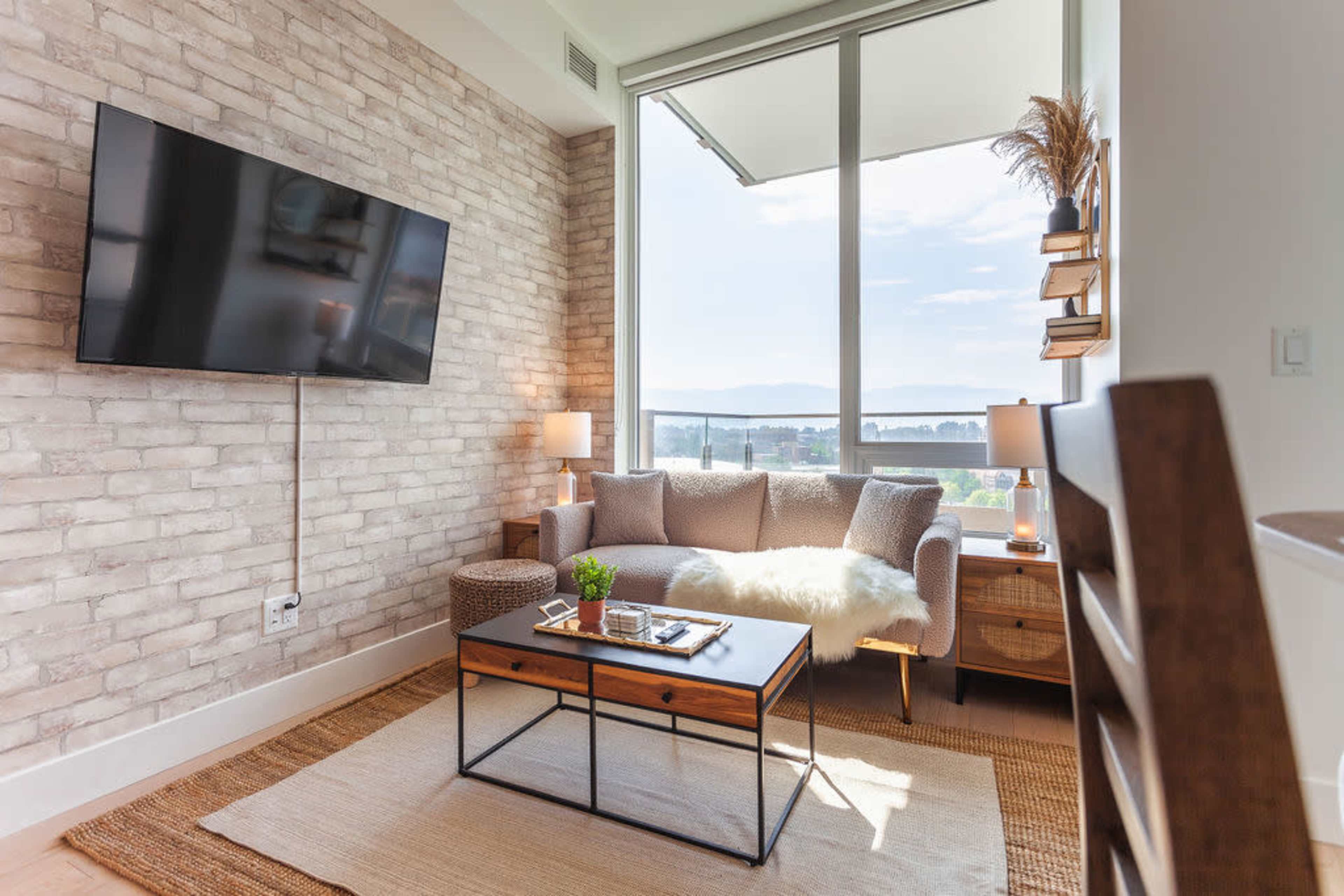 The image shows a cozy living room with a gray couch, a wood coffee table, a mounted television, and large windows providing a scenic view.