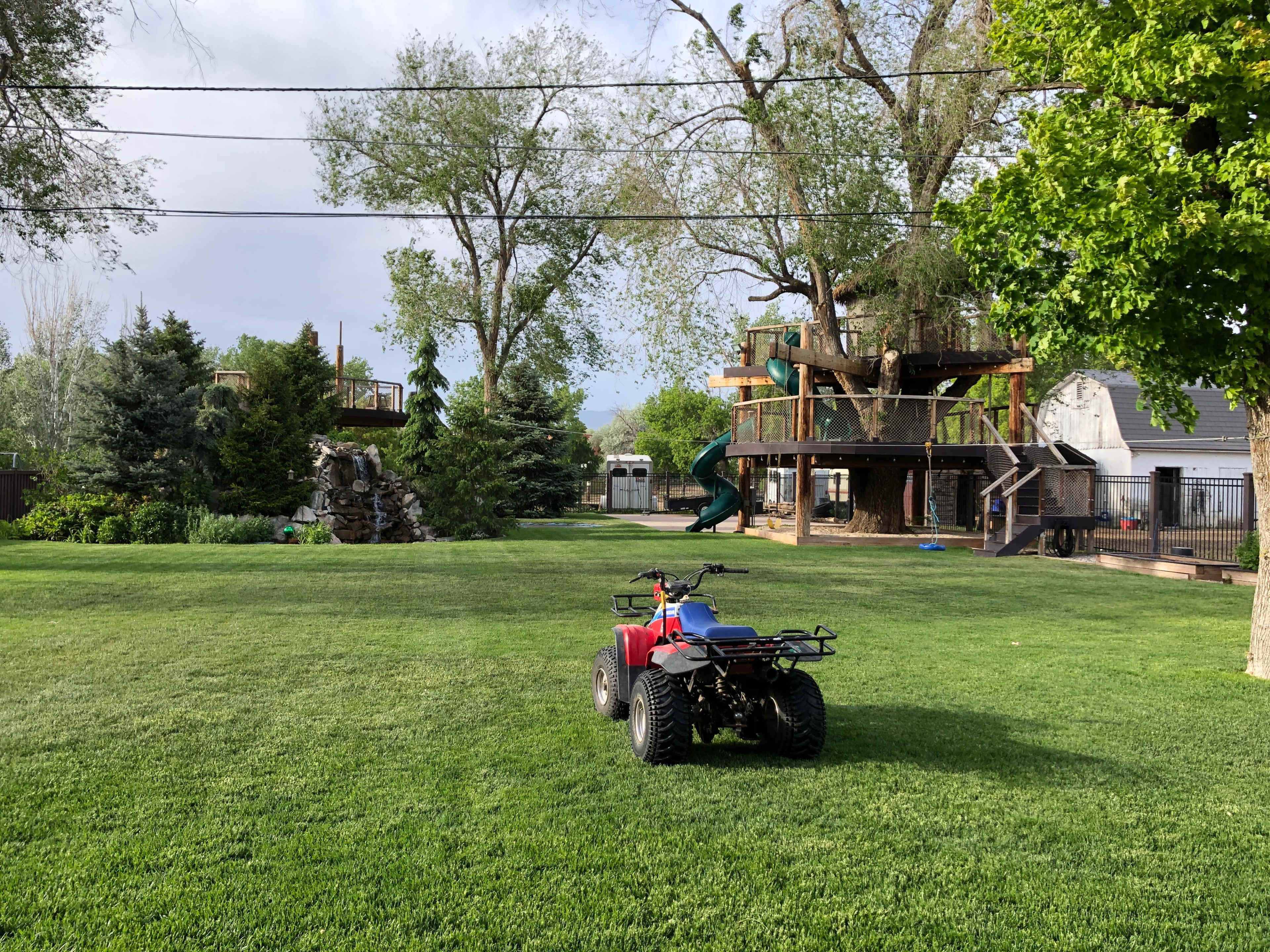 A red ATV is parked on a green lawn surrounded by trees, play structures, and a rock formation.