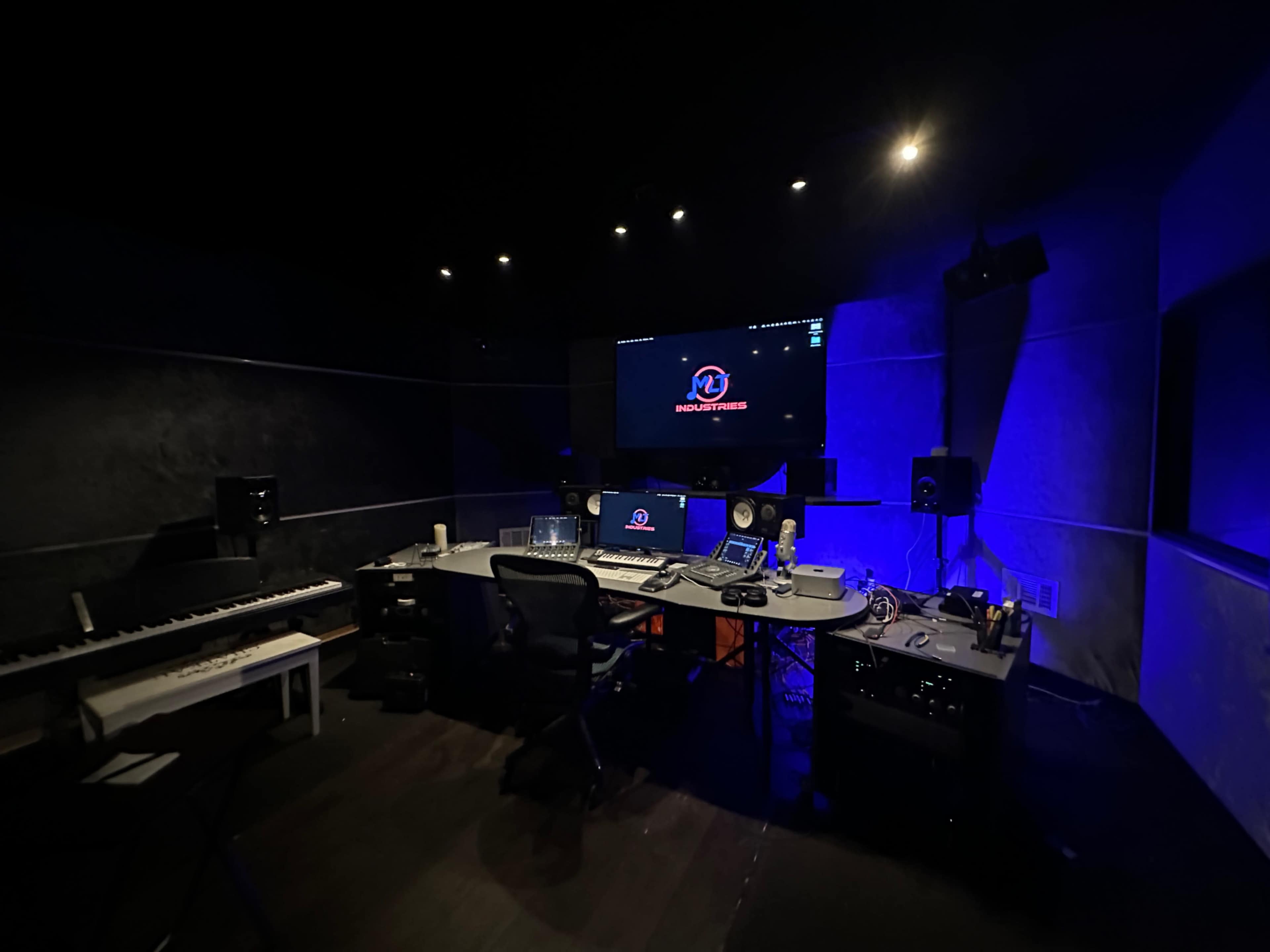 The image shows a dimly lit recording studio with a central desk, multiple screens, and audio equipment along with a keyboard.