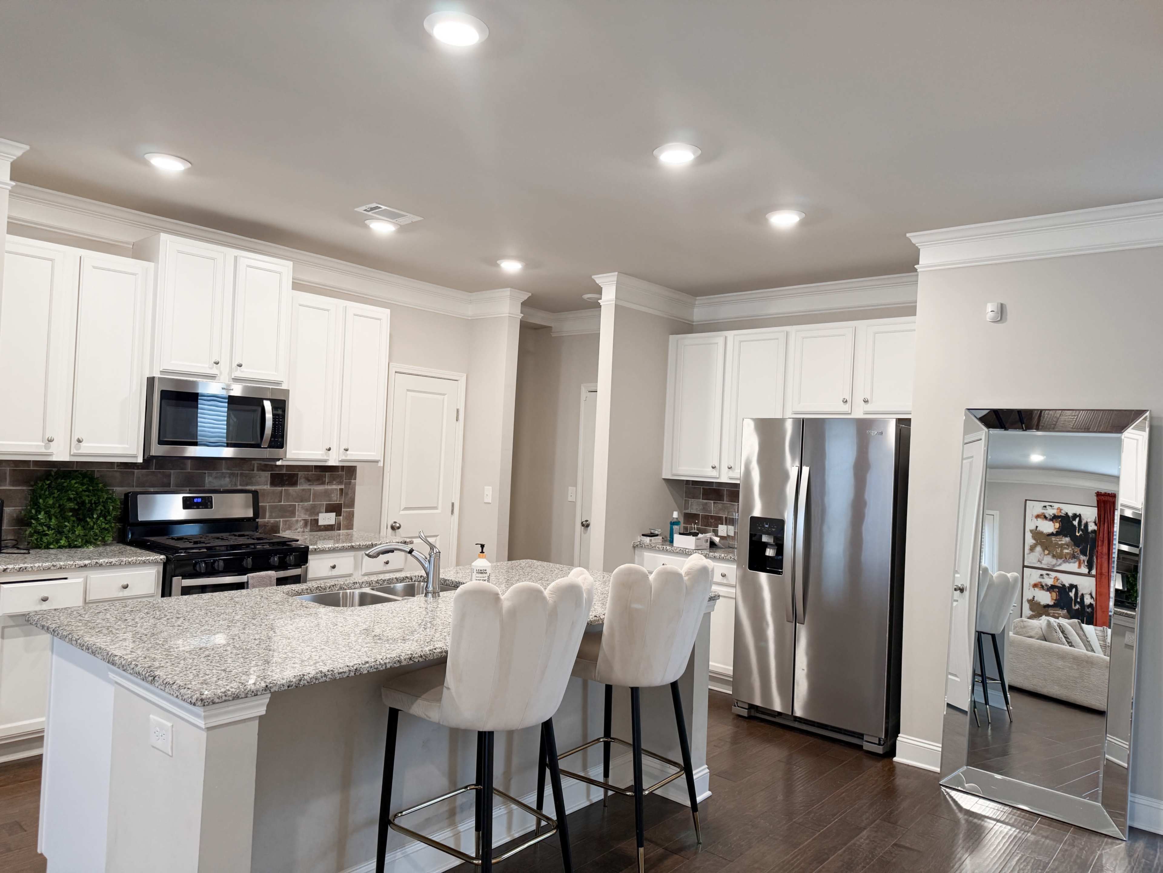 The image shows a modern kitchen with white cabinetry, a granite countertop island, stainless steel appliances, and bar stools.