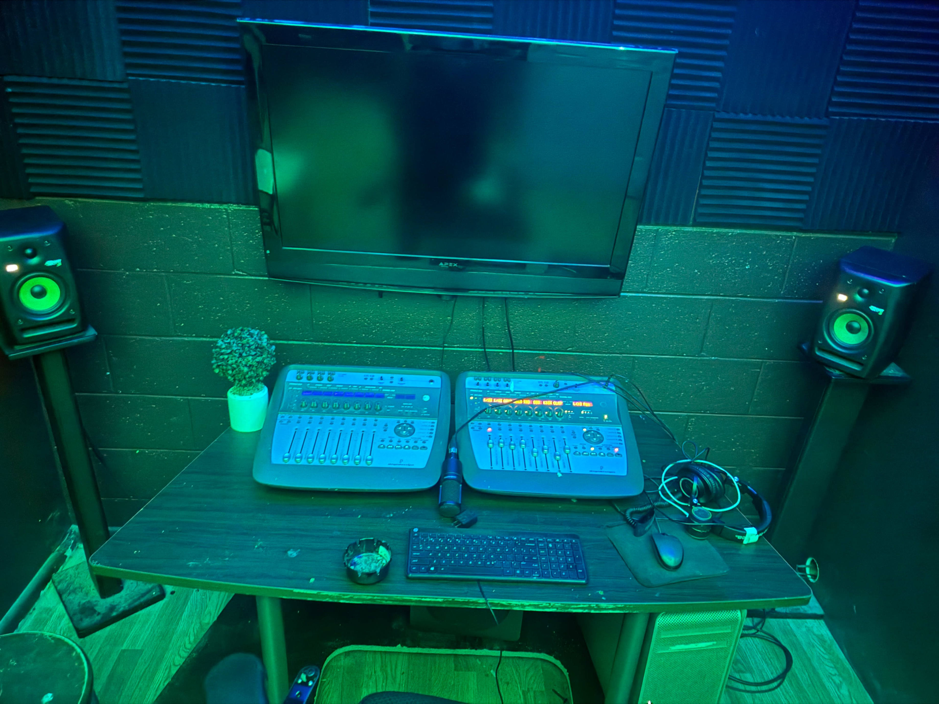 The image shows a small recording studio with a desktop mixing console, a computer monitor, and two studio speakers on either side, all set against a textured wall.