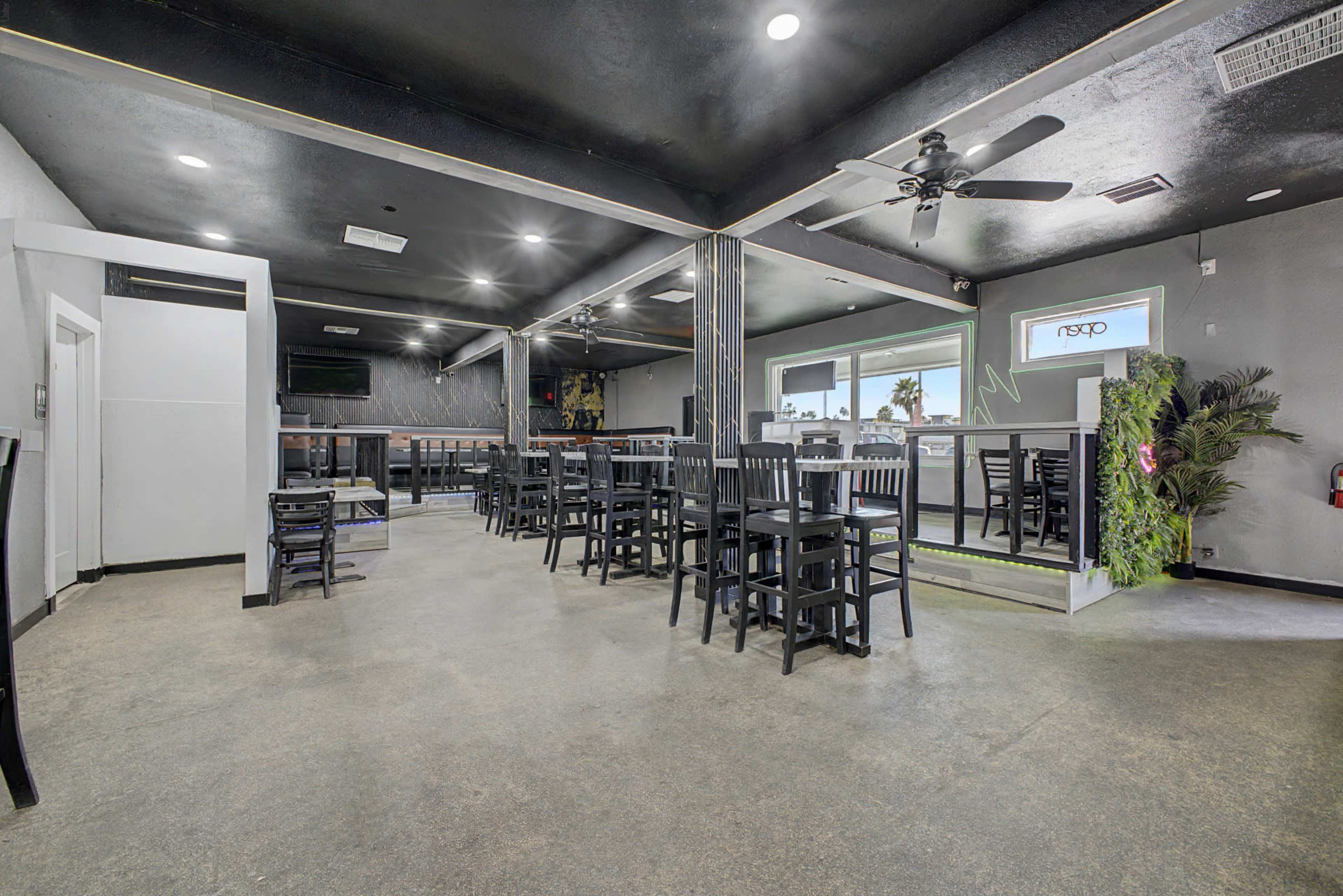 The image shows a spacious indoor dining area featuring several black wooden tables and chairs, a bar at the back, and large windows illuminated by natural light.