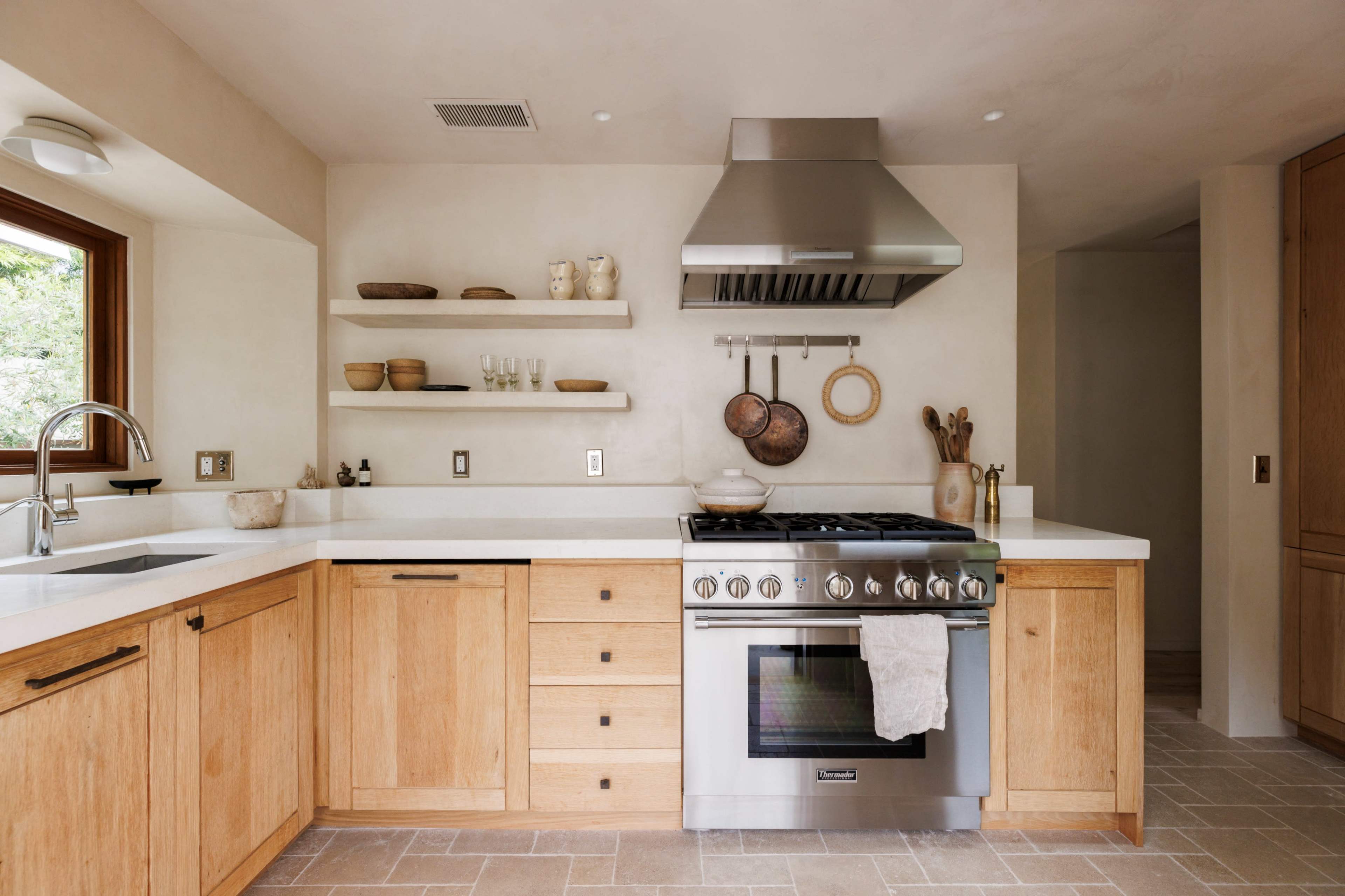 A modern kitchen features wooden cabinetry, a stainless steel range hood, and open shelving displaying ceramic dishes and utensils.
