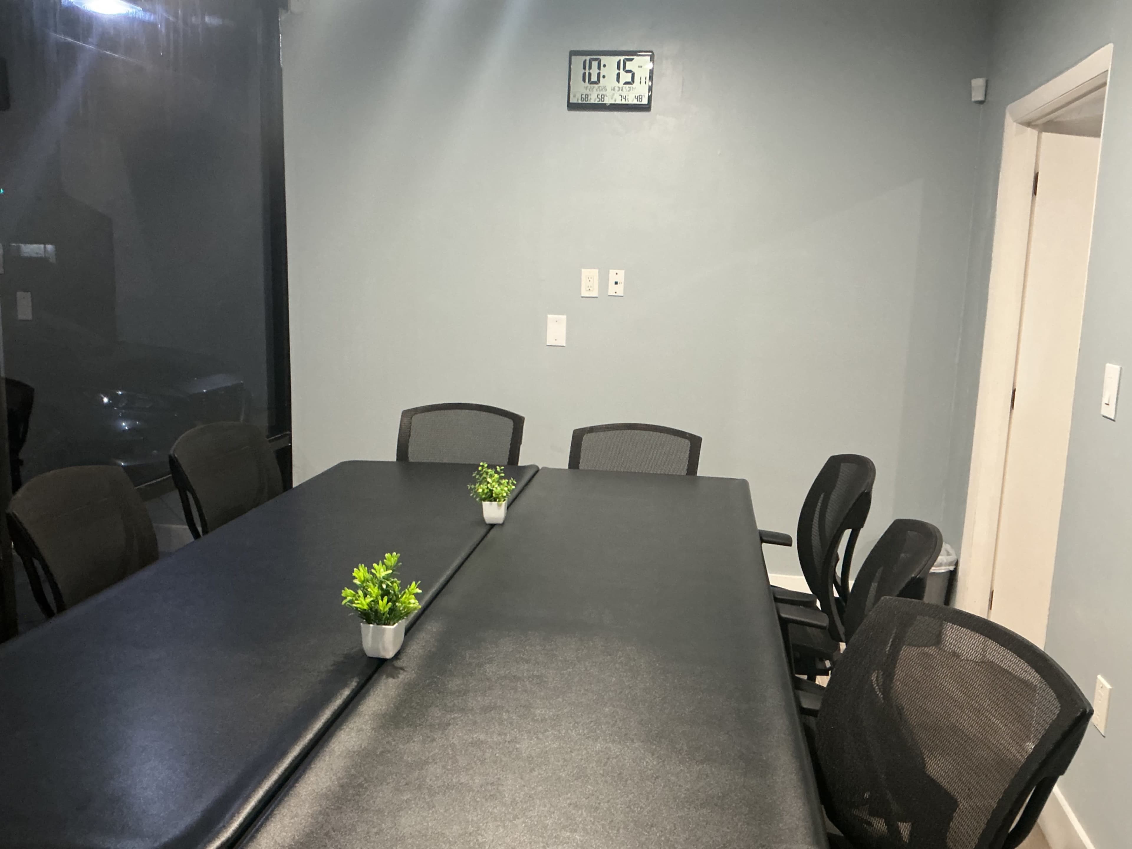 A conference room features a rectangular black table surrounded by six mesh chairs, with small green plants placed in the center and a clock displaying 10:15 on the wall.