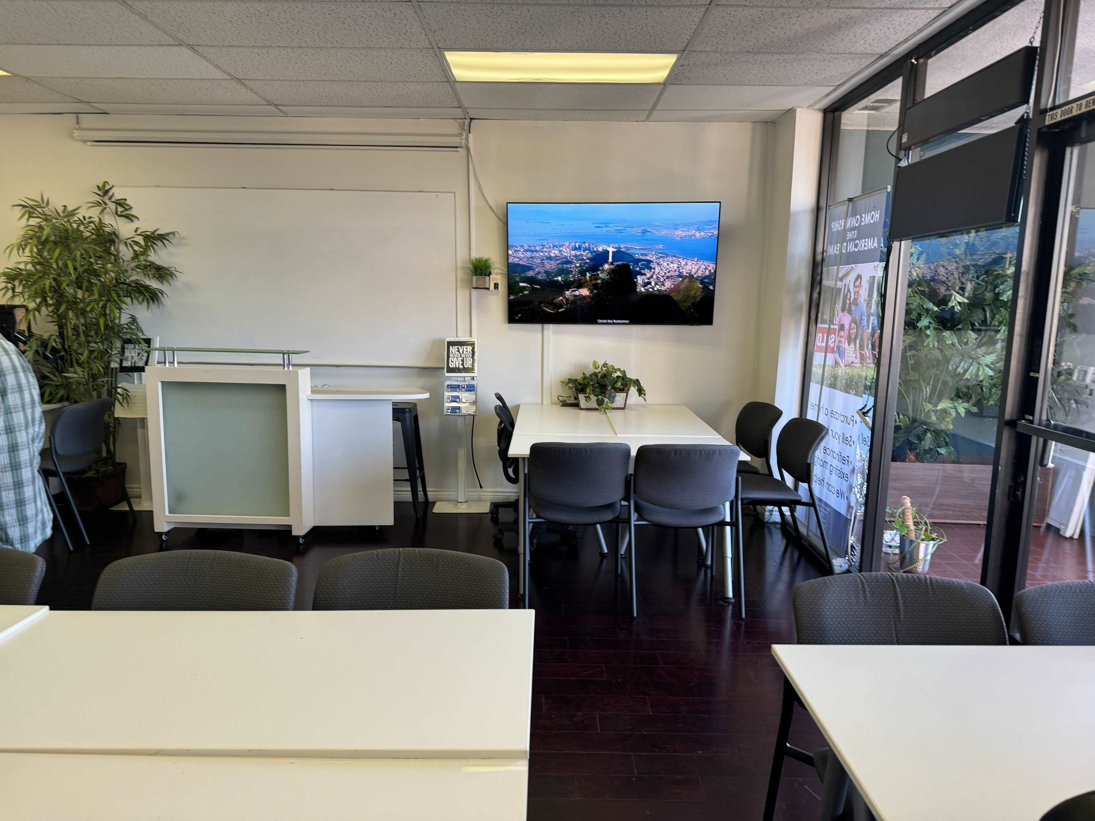 A well-lit classroom or meeting space with white tables, chairs, a large TV screen displaying a city view, and plants in the background.