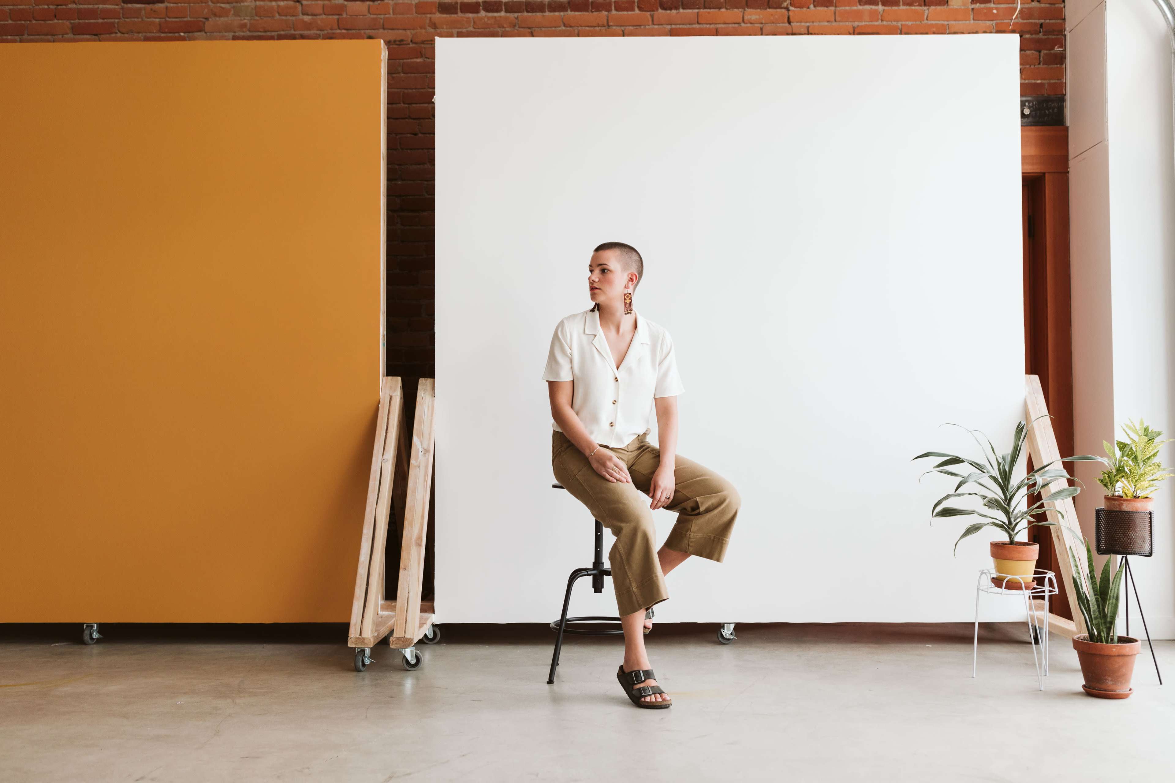 A person sits on a chair in a brightly lit room with two large backdrop panels, one orange and one white, while plants are placed nearby.
