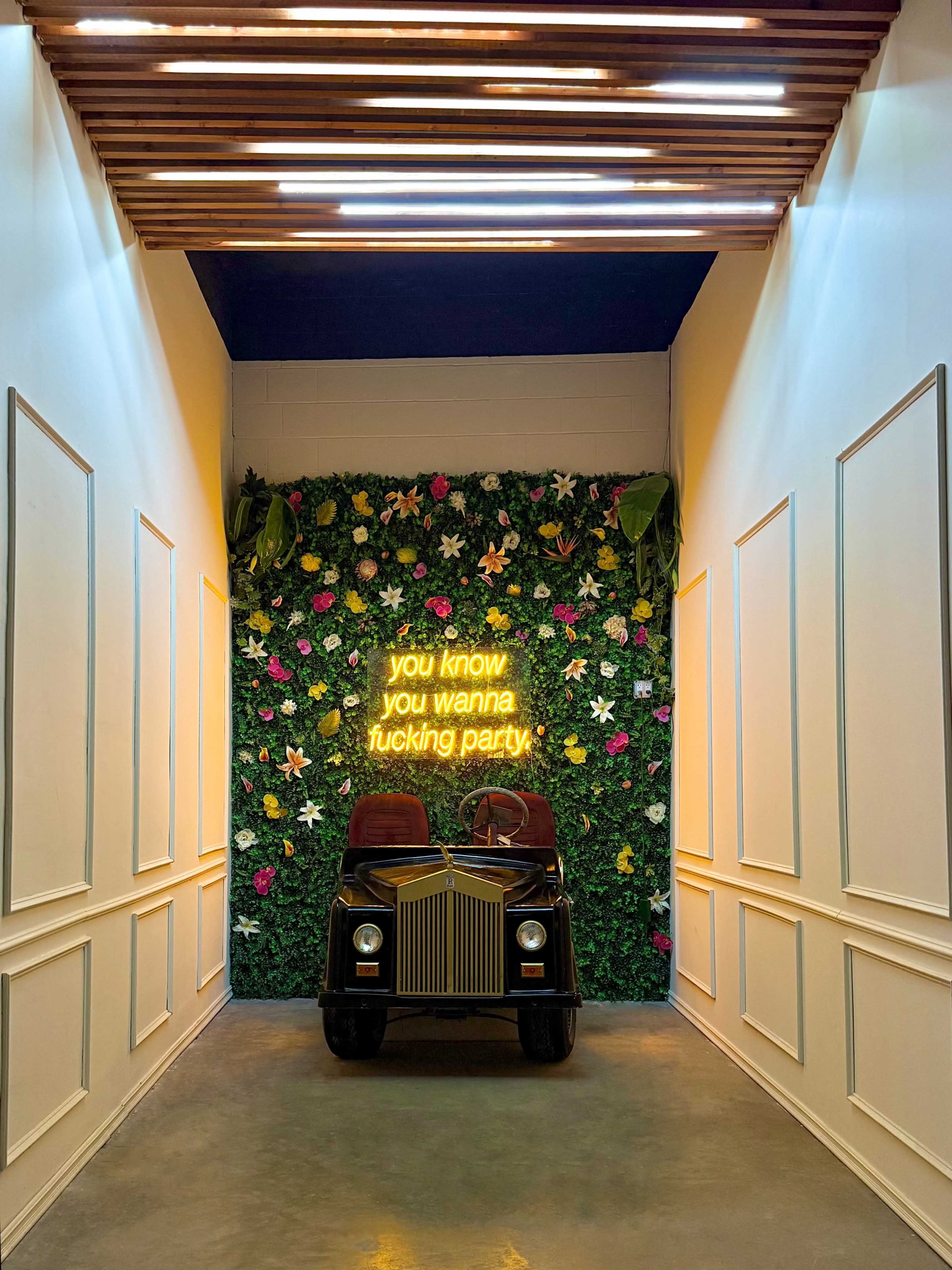 A vintage car is positioned in a brightly lit hallway adorned with a green floral wall and a neon sign that reads, "you know you wanna fucking party."