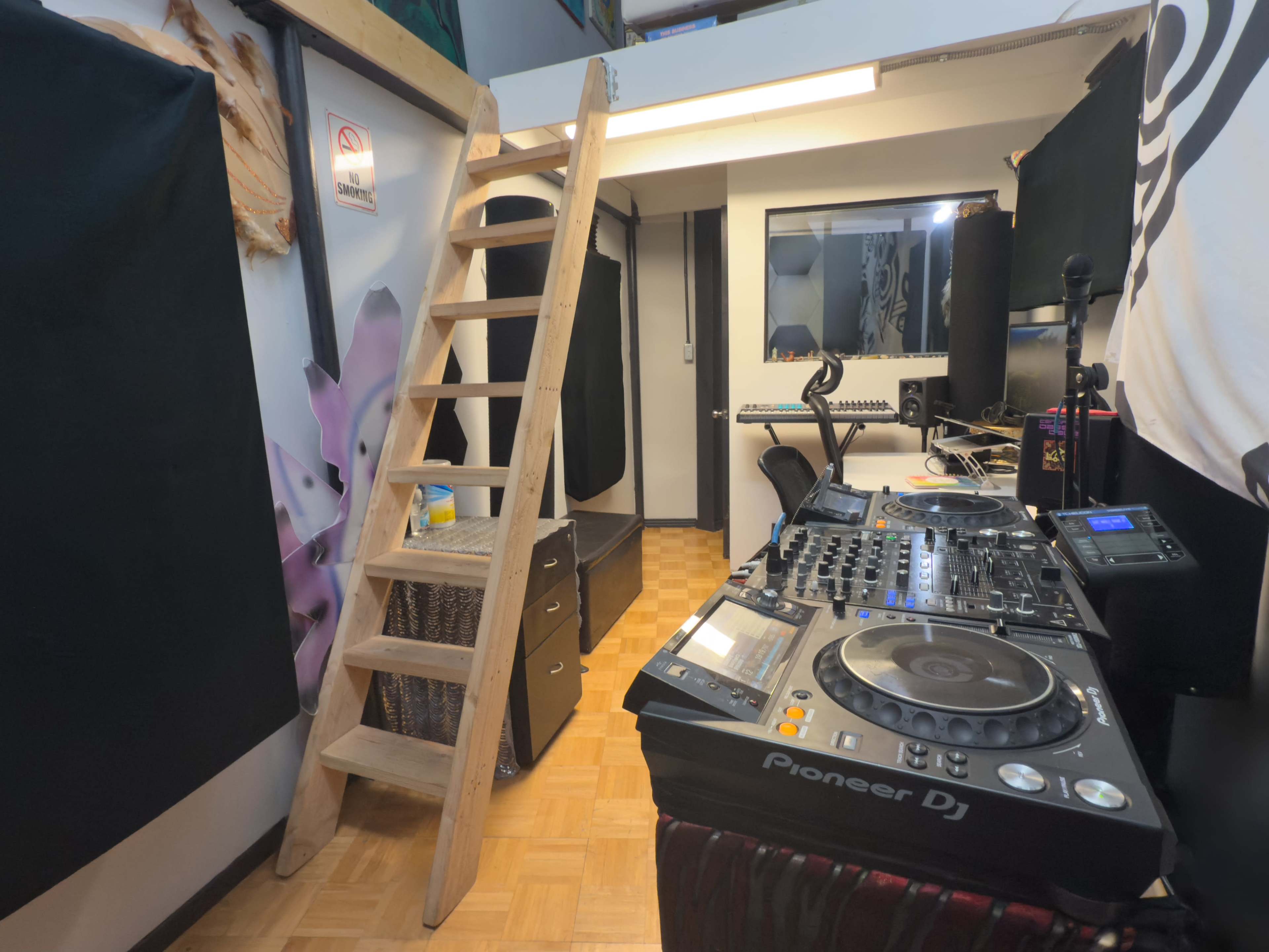 The image shows a small DJ room with a wooden ladder, a DJ setup featuring Pioneer equipment, and a mirror surrounded by black panels.