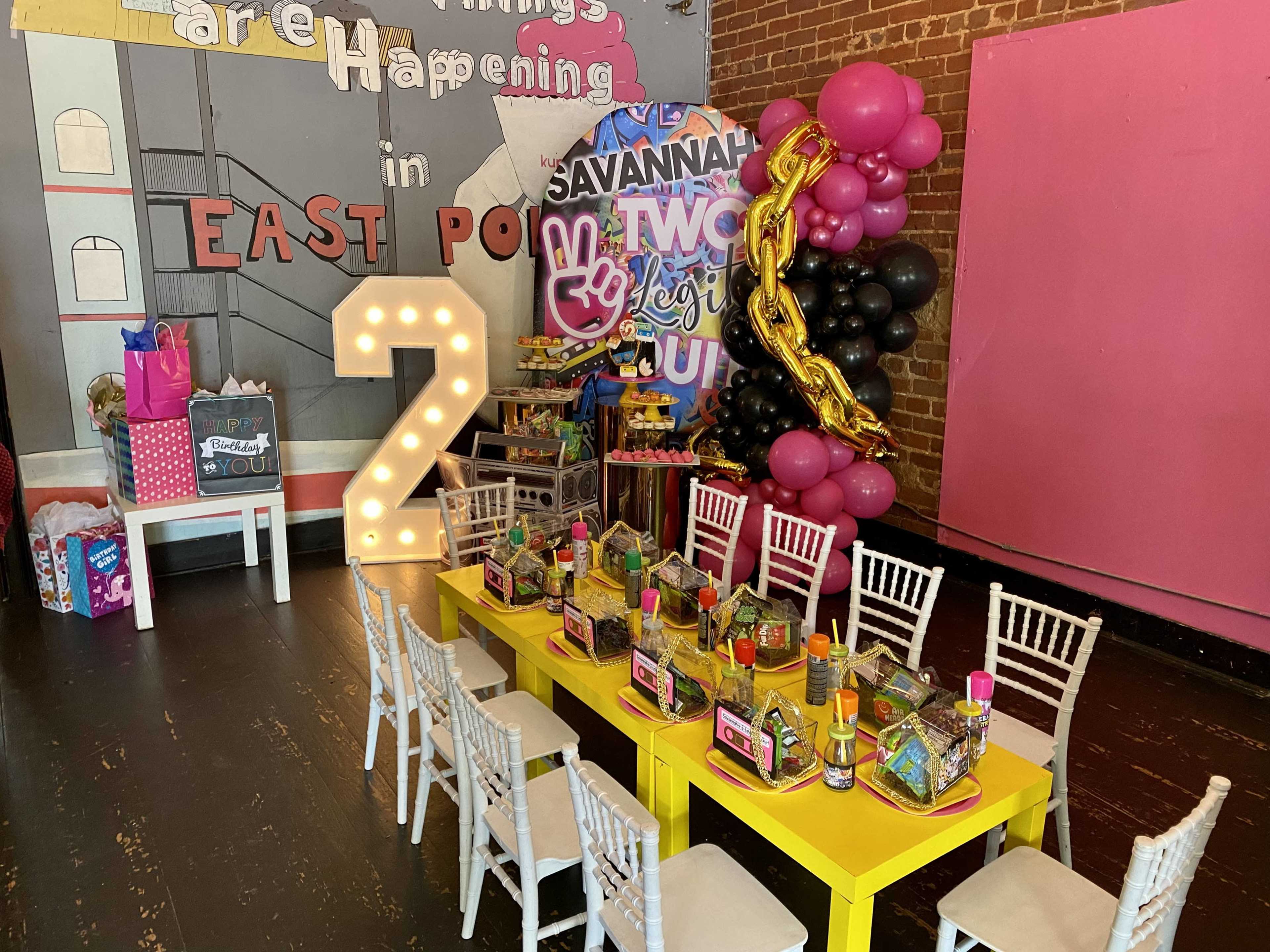 A festive party setting features a yellow table arranged with colorful party favors and decorations, including a large pink and black balloon display and a prominent "2" marquee light, set against a brick wall.