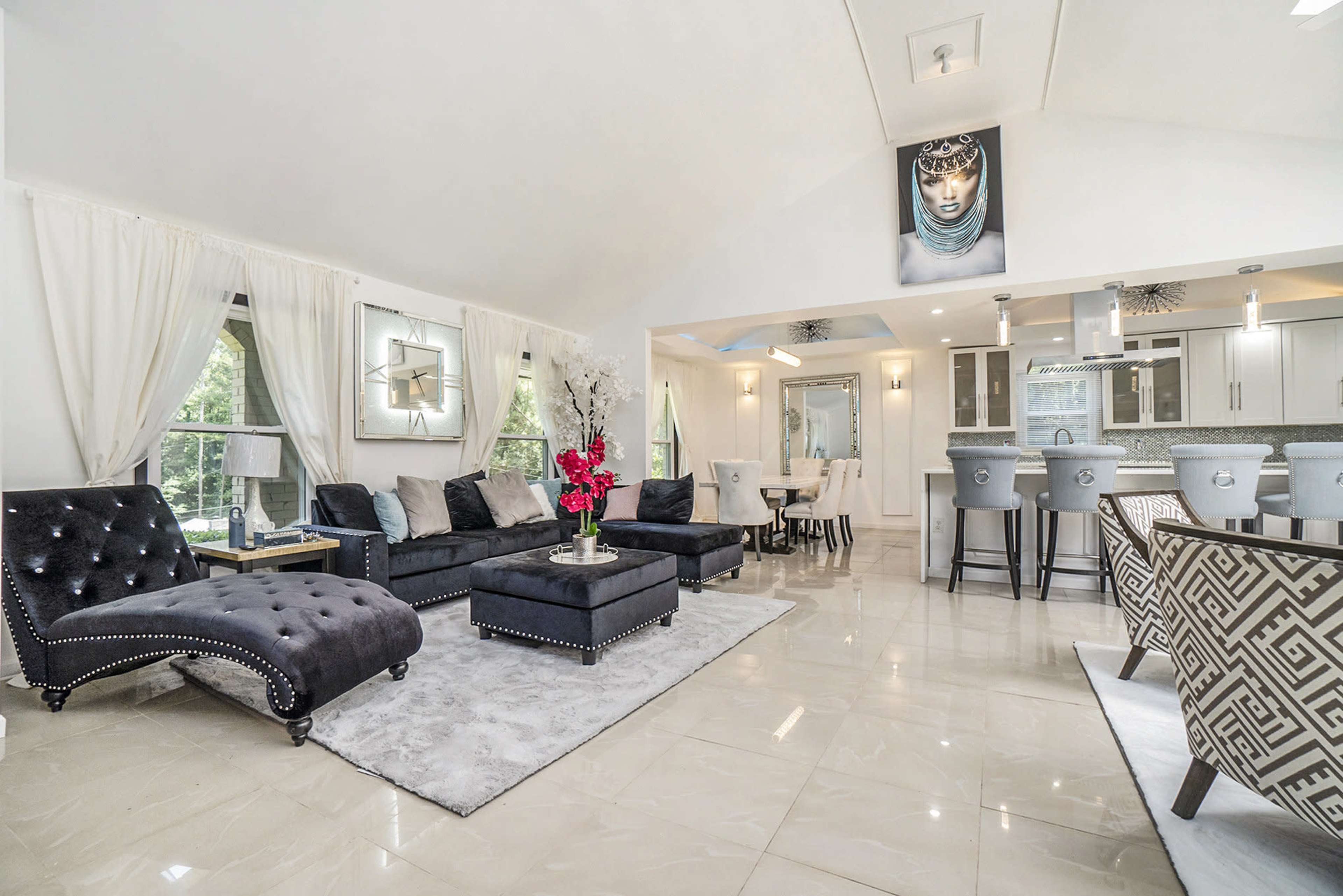 The image shows a modern living room with a black couch and chaise lounge, a light-colored rug, and a dining area featuring elegant chairs and a kitchen in the background.