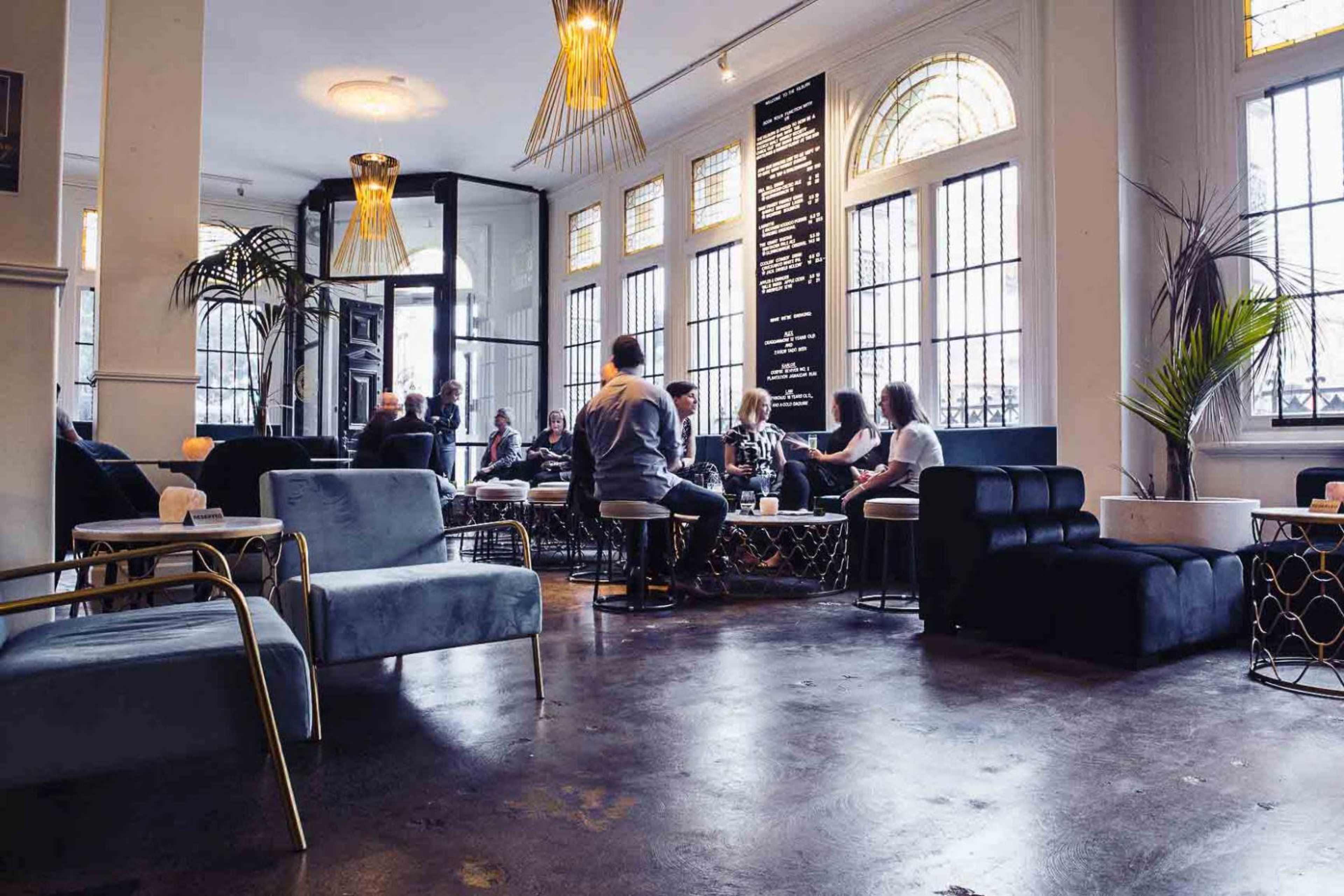 A modern café interior features seating arrangements with people socializing, large windows allowing natural light, and decorative lighting fixtures.
