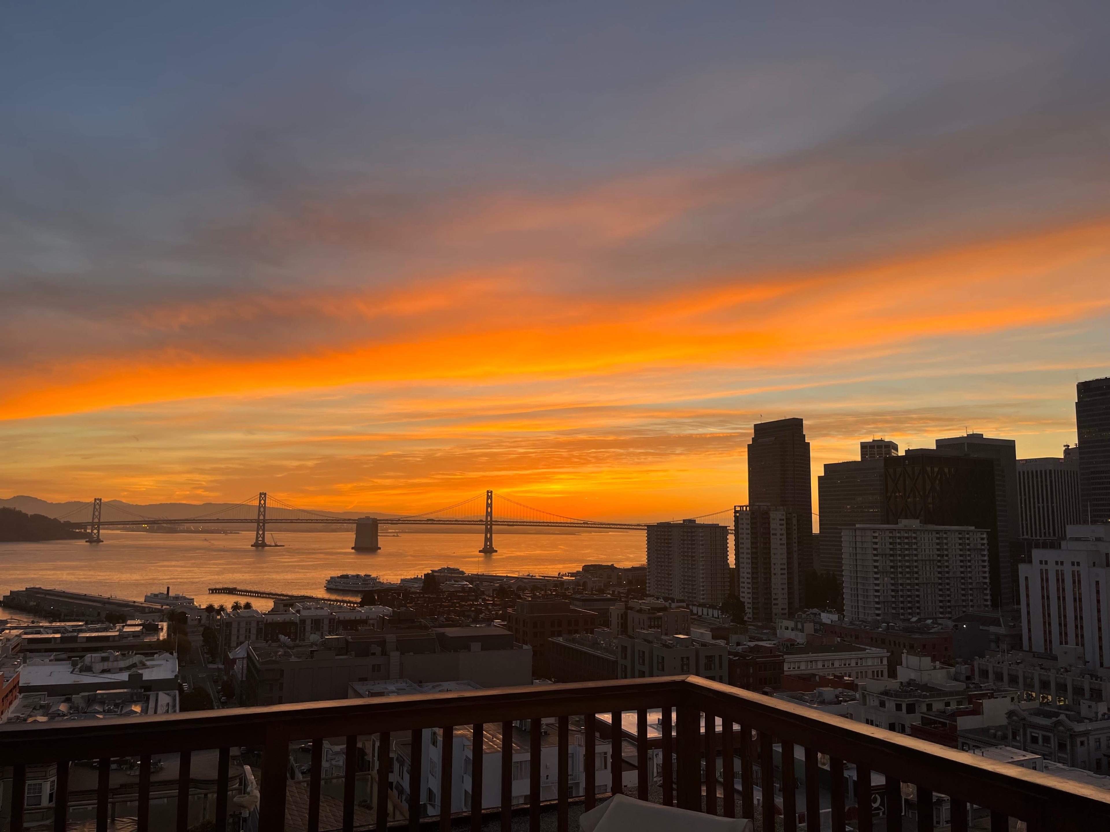 A vibrant sunset over the San Francisco Bay, with the Bay Bridge visible and the skyline of buildings in the foreground.