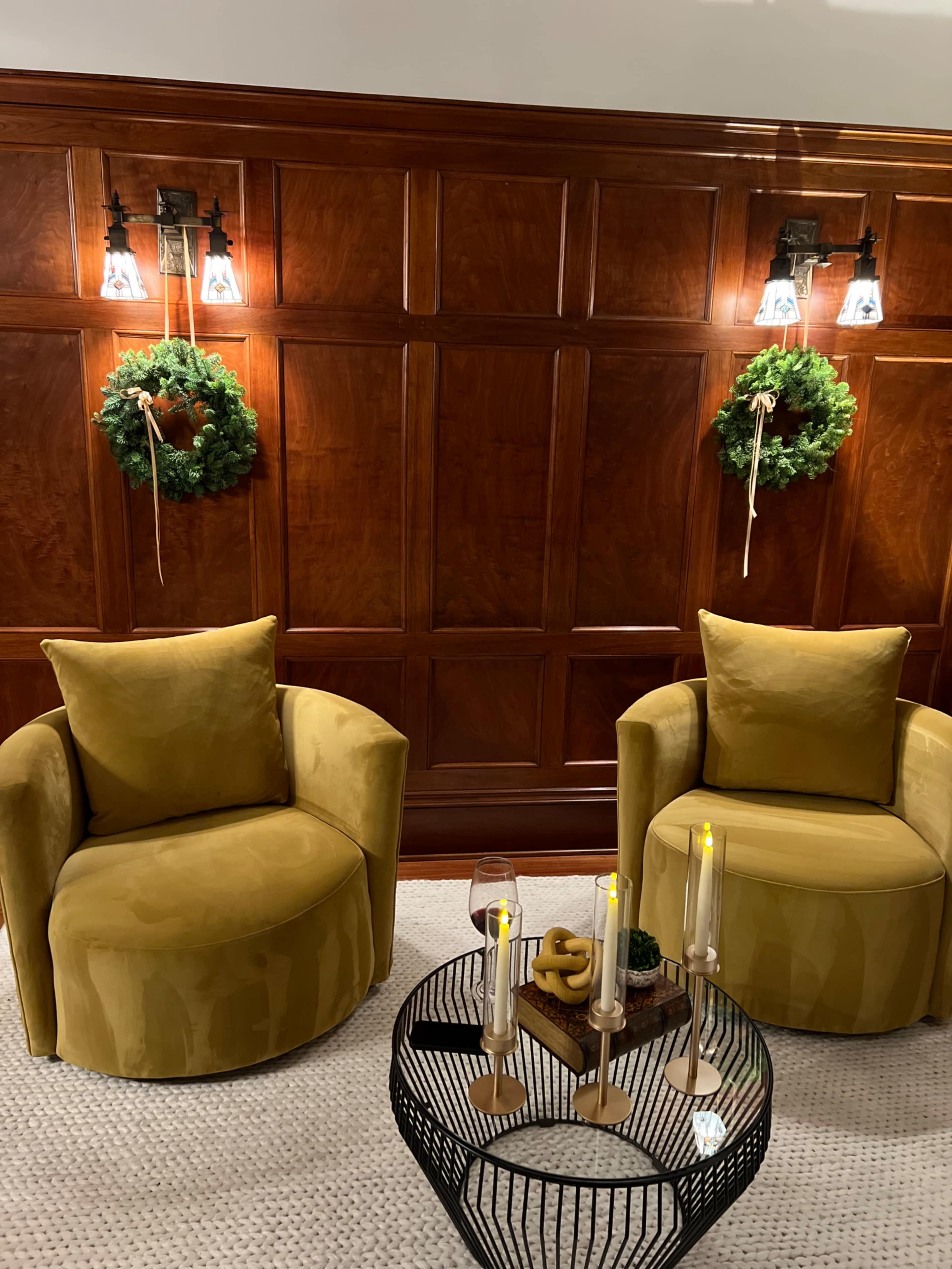 The image shows two round, mustard-colored chairs positioned near a decorative wooden wall, adorned with two green wreaths and lit candles on a circular black metal coffee table.