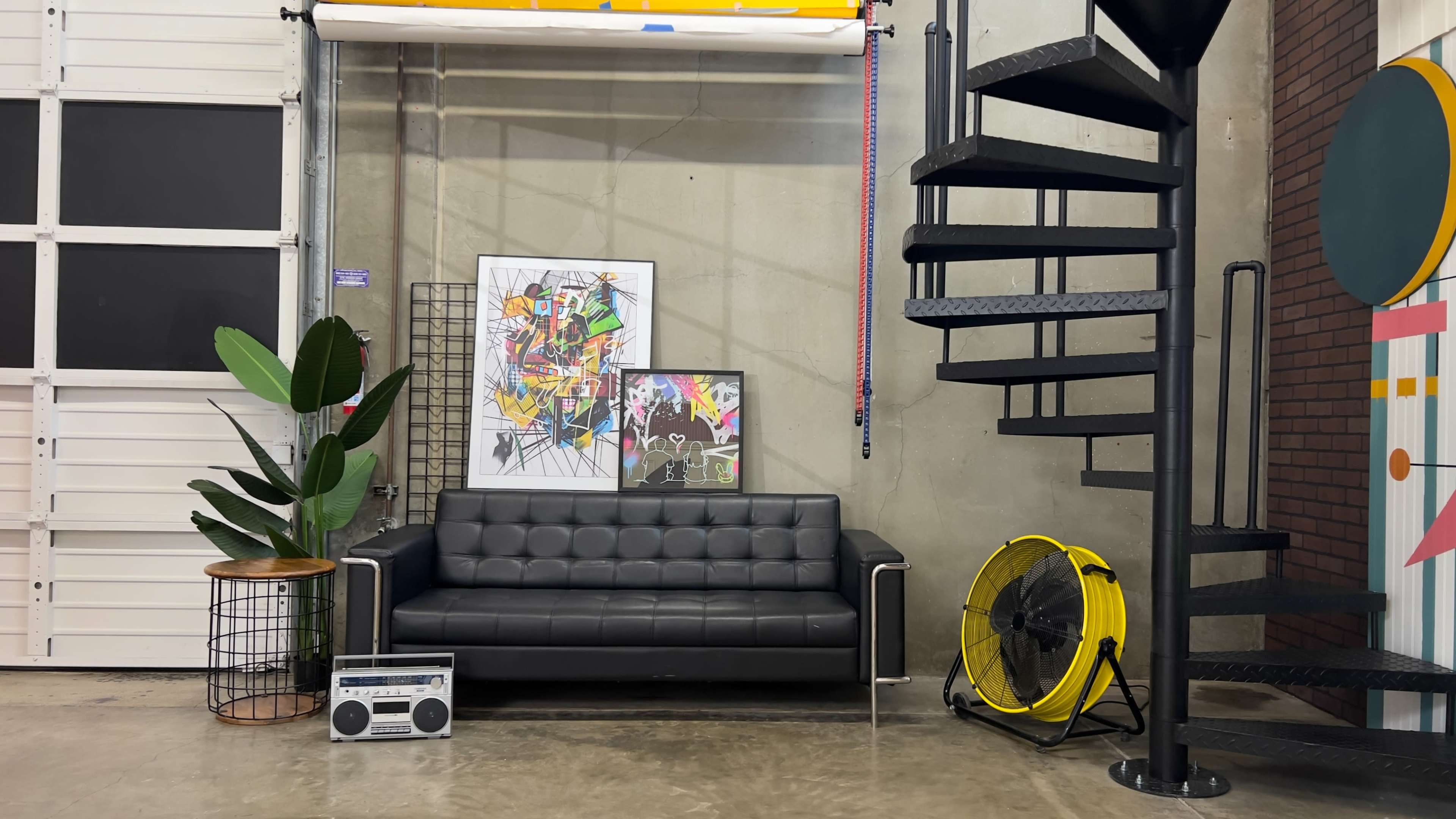 A black couch is positioned against a textured wall, accompanied by two framed artworks, a potted plant, a vintage boombox, and a large yellow fan near a spiral staircase.